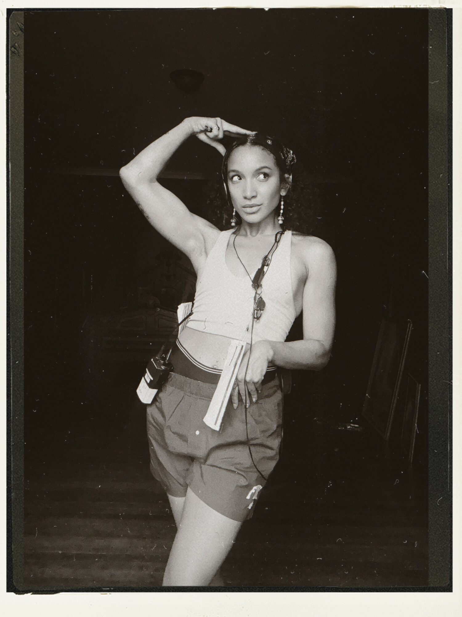 Young woman with short hair, wearing a tank top and shorts, holding a book and a camera, standing indoors.