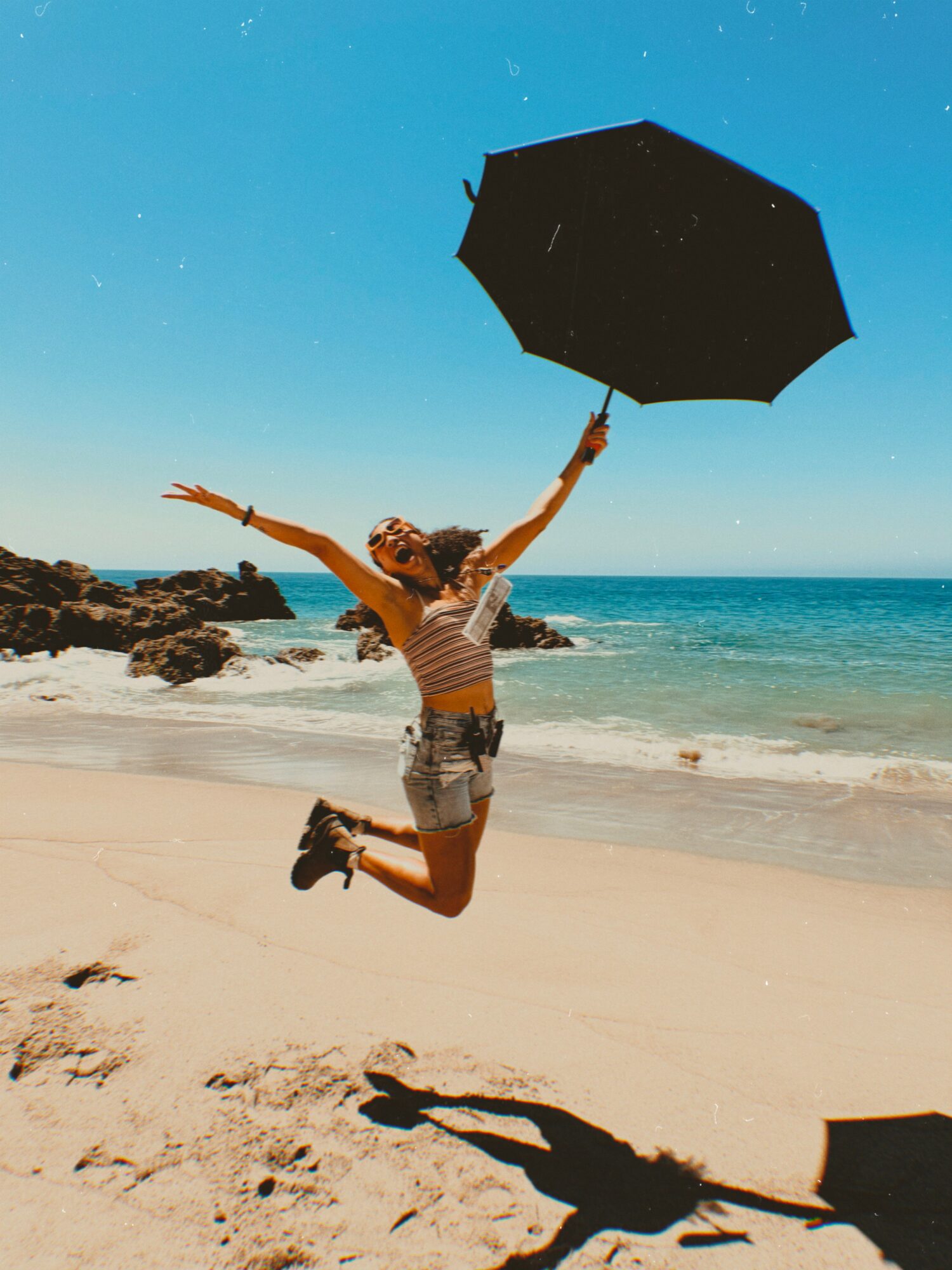 Person jumping on beach holding an umbrella, with ocean and sky in background.