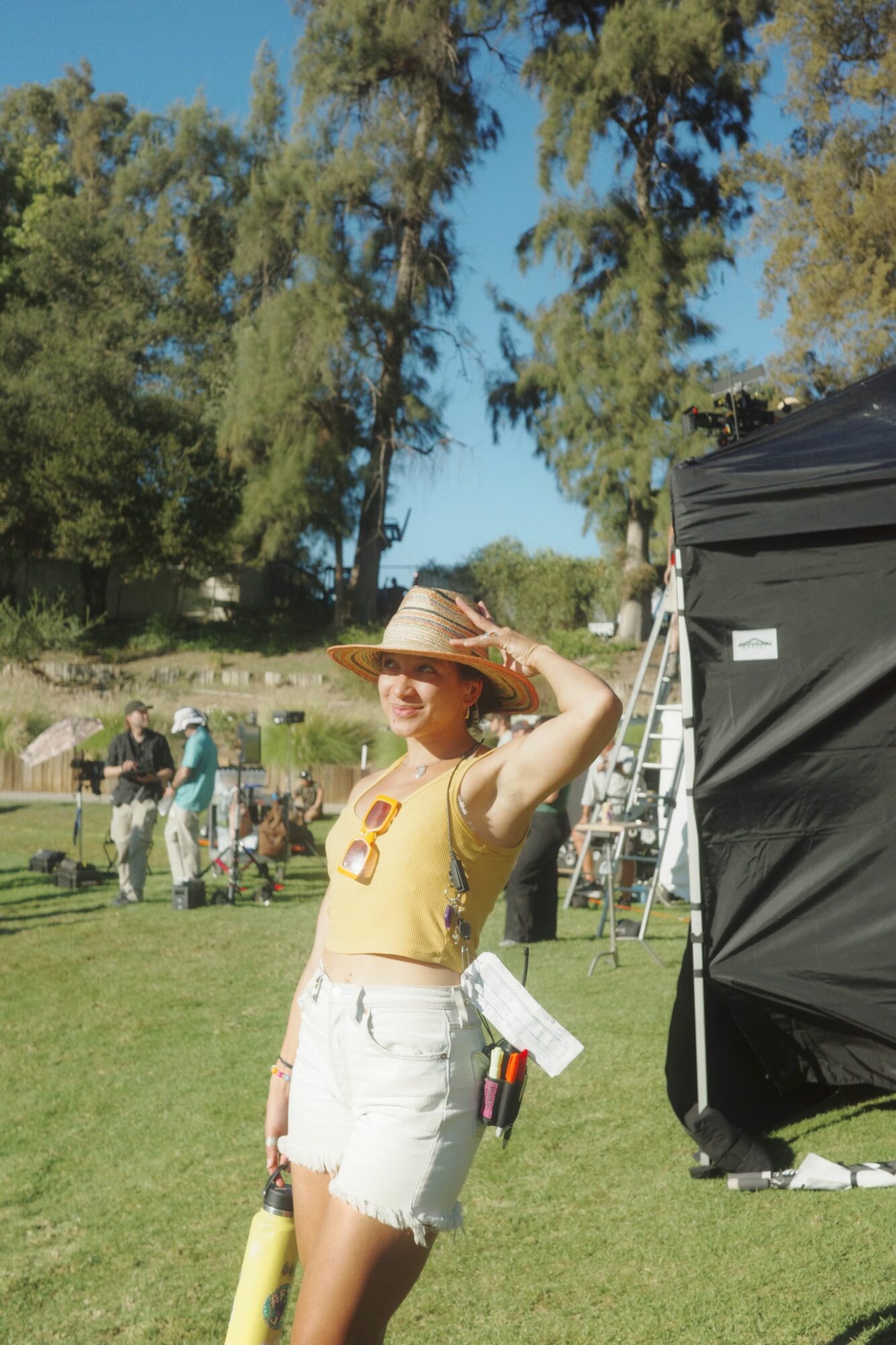 Person in yellow top and white shorts holding a hat, standing on grass with trees and people in background.