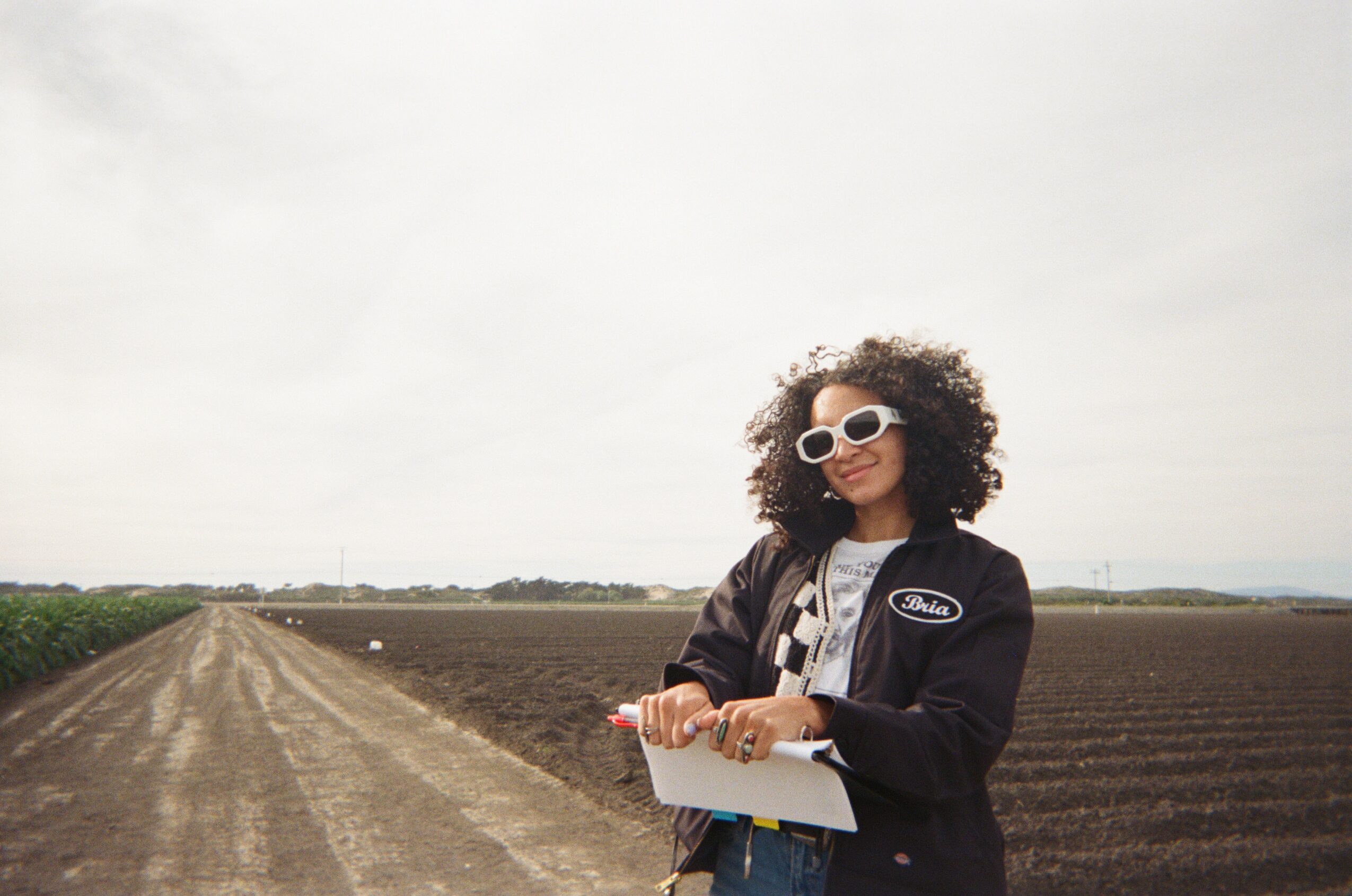 Person with curly hair wearing sunglasses and a jacket holding a clipboard stands outdoors near a dirt road and farmland.