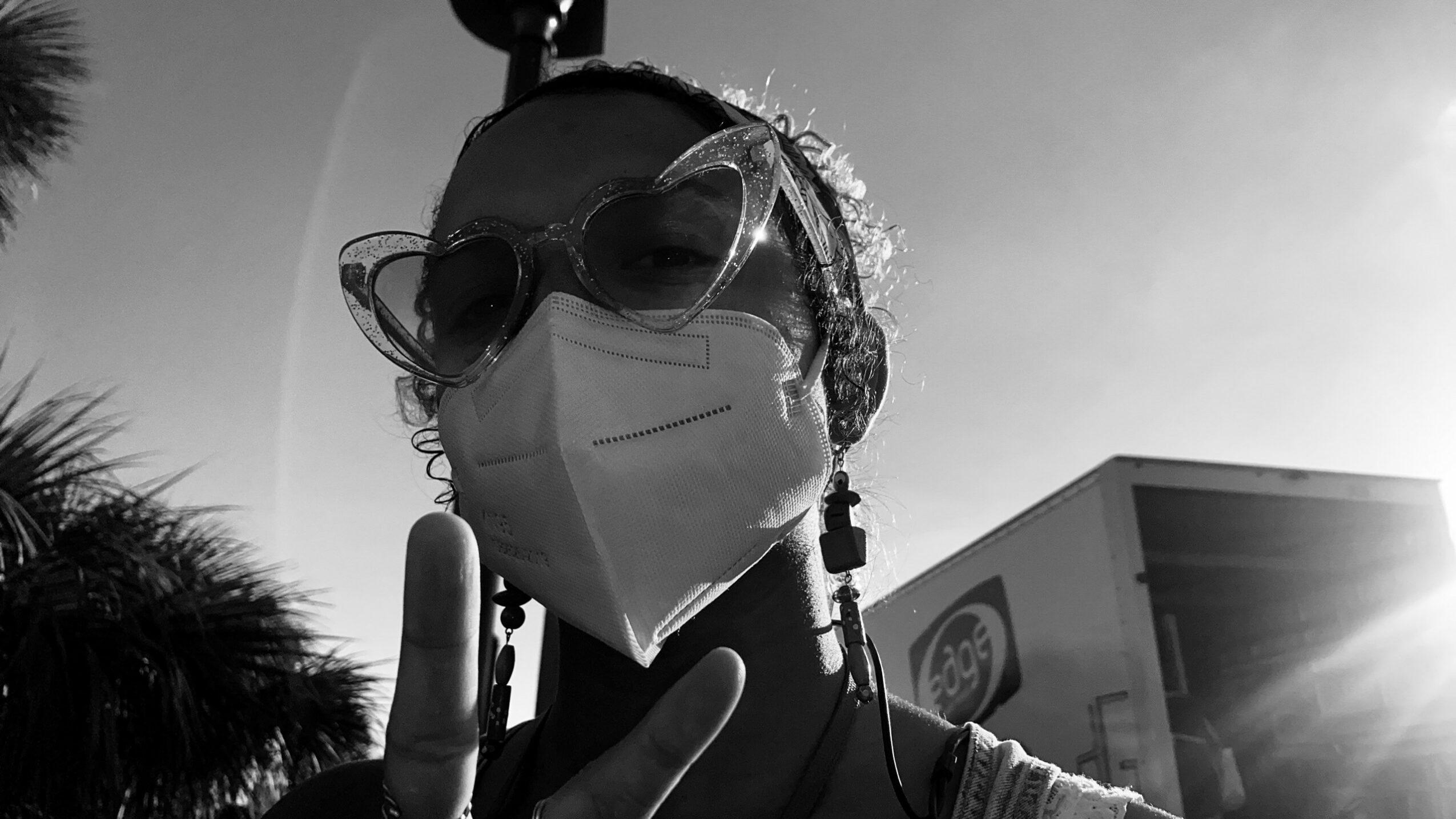 Person wearing sunglasses and a face mask making a peace sign outdoors, with sunlight and a building in background.