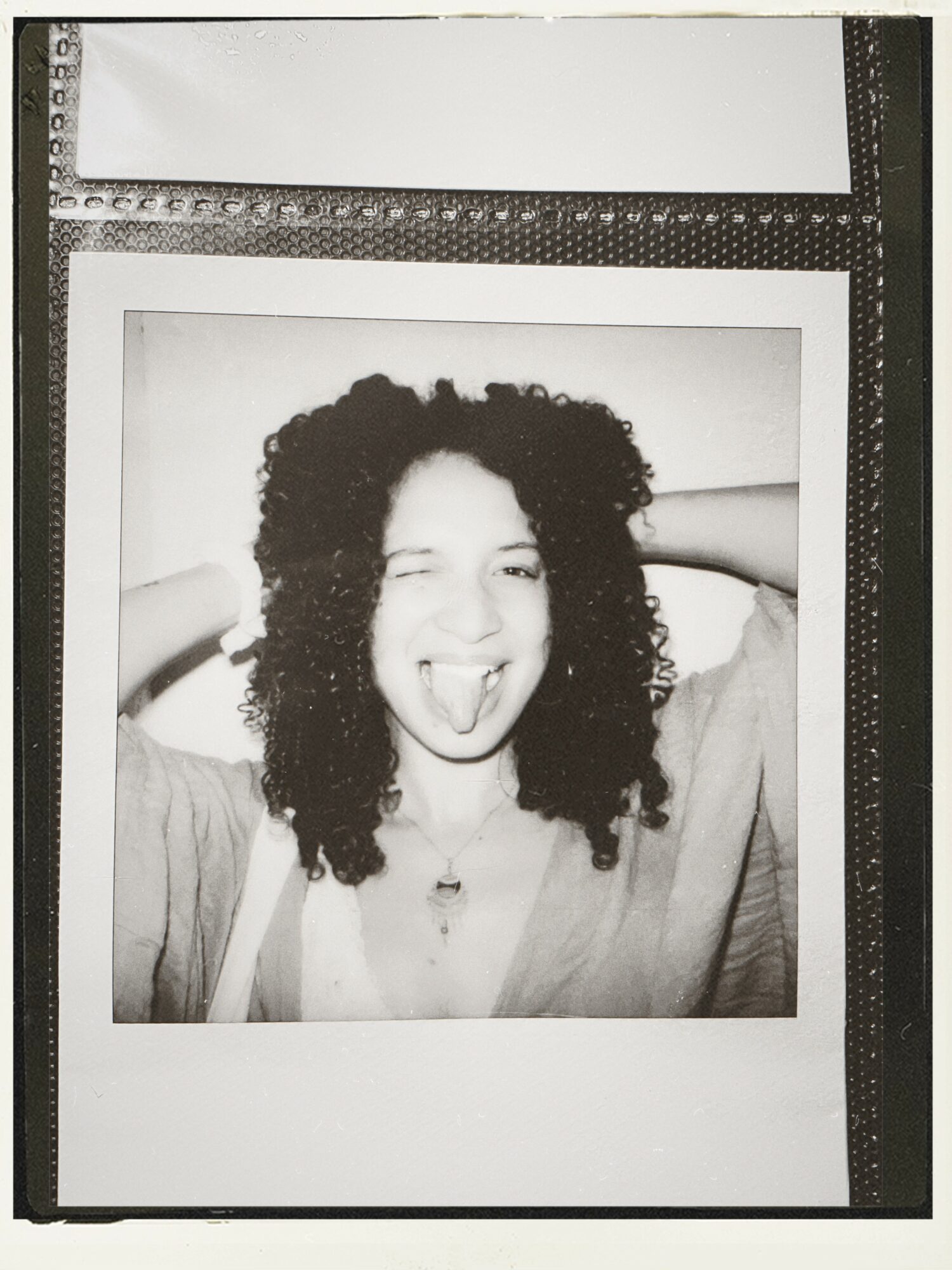 Person with curly hair sticking out tongue, smiling, holding hands behind head, in a Polaroid-style photo, black and white.