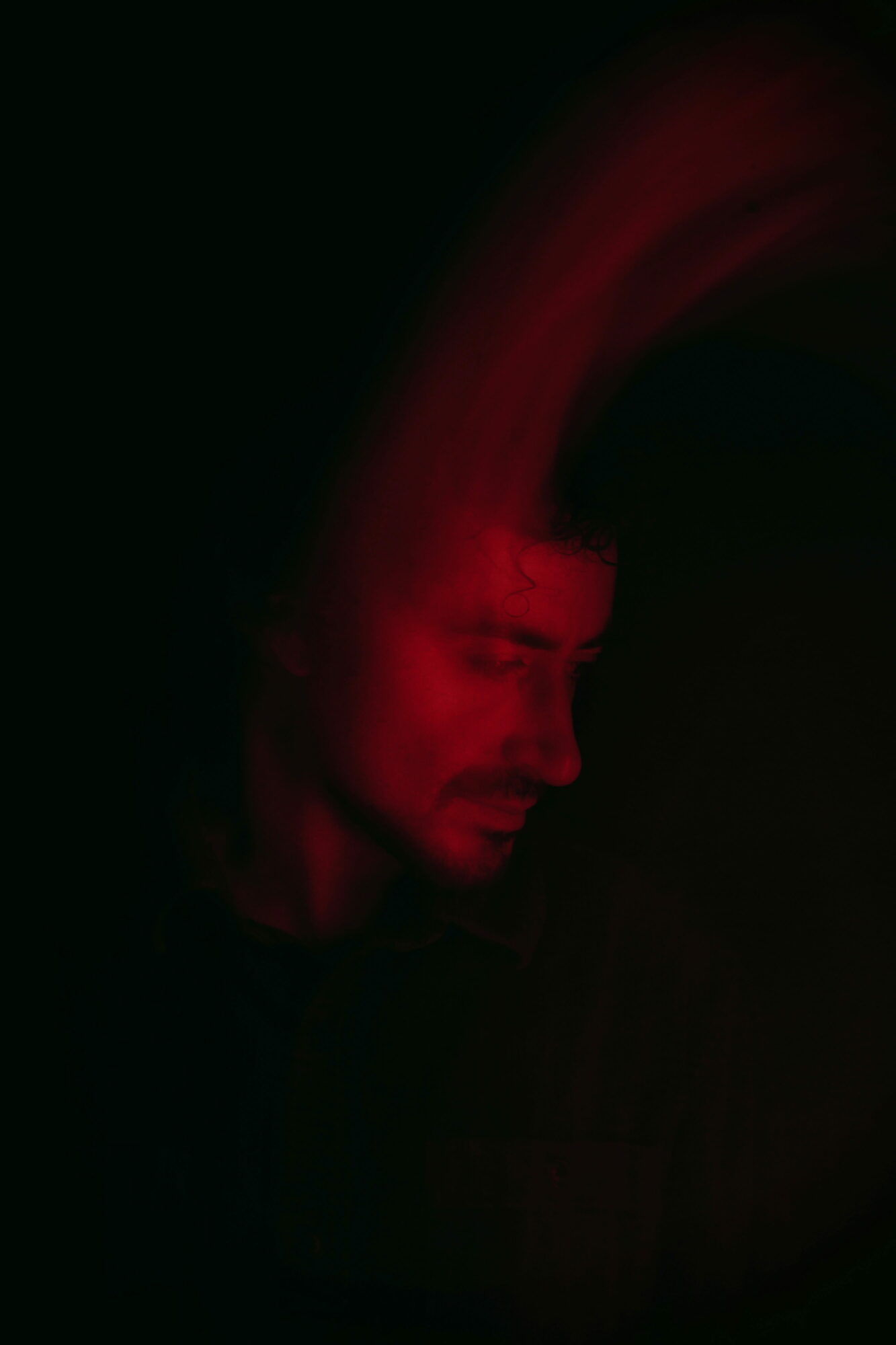 Person with long hair illuminated by red light, head tilted downward, dark background.