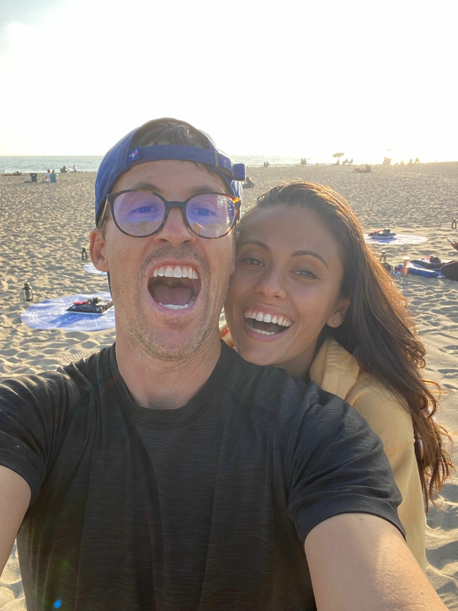Two people smiling and taking a selfie at the beach with sand and water in the background.