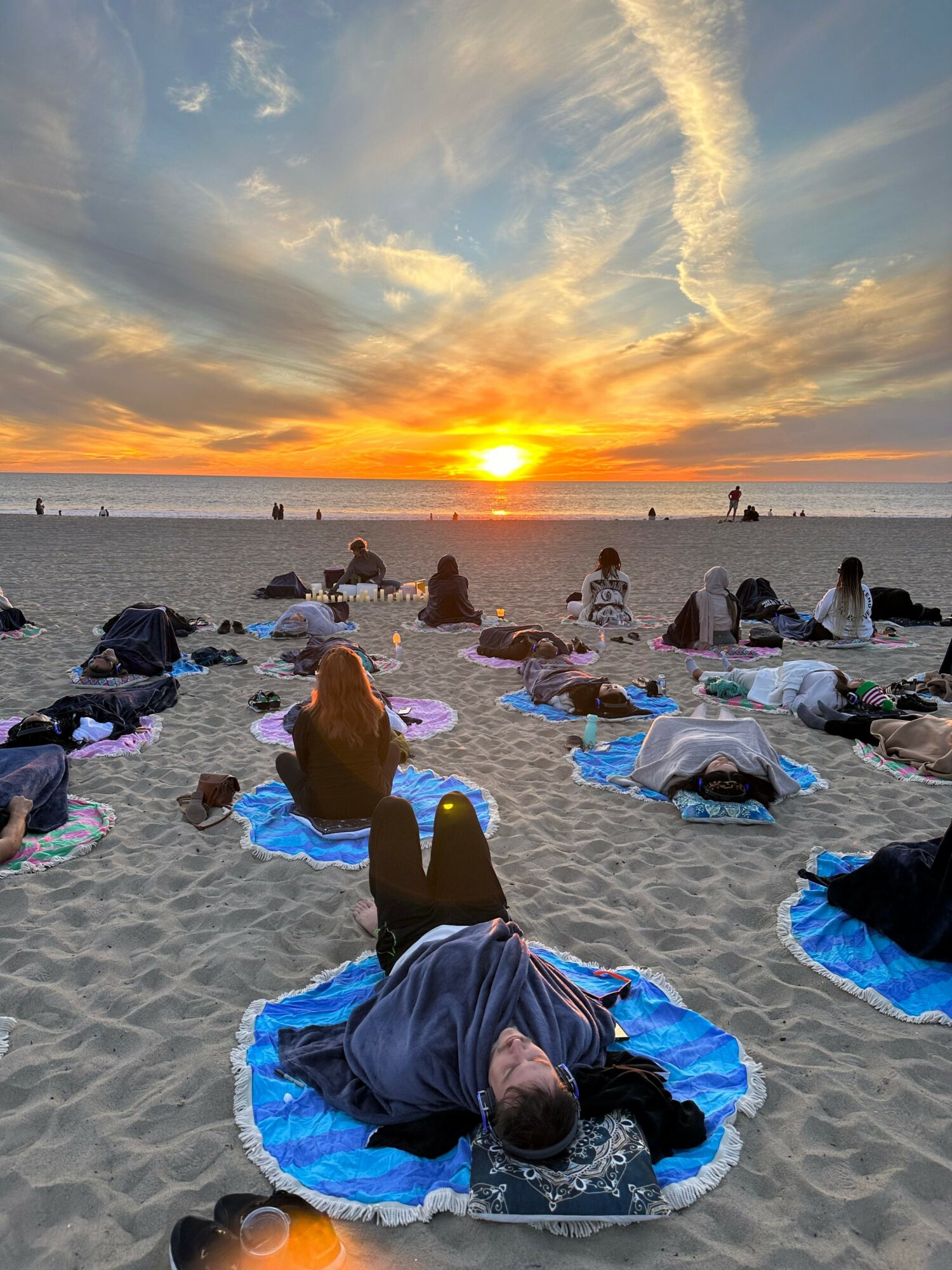 People sitting on the beach during sunset, some on towels, with a person lying down in the foreground, sky with clouds and sun.