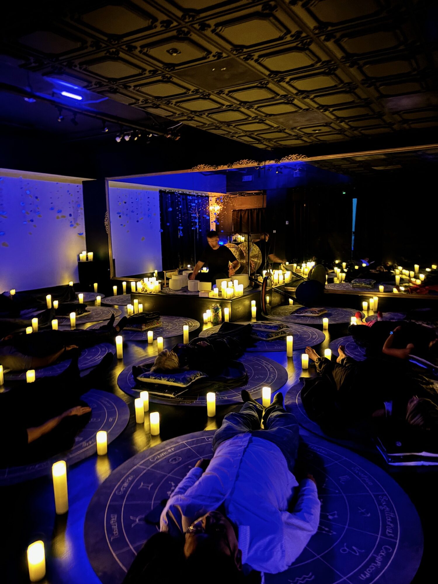 People lying on the floor in a dimly lit room with candles and a projection screen, possibly during a meditation or event.