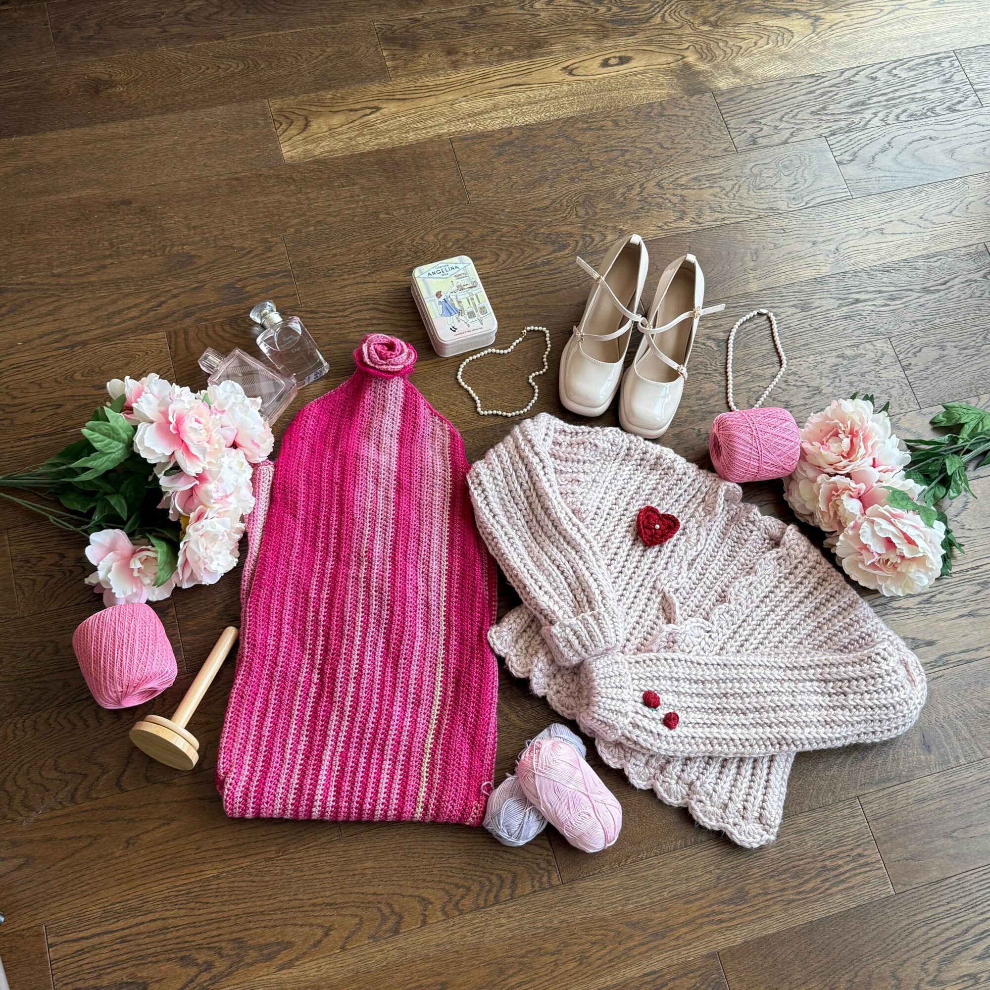 Pink knit hat, cream sweater, white shoes, pink purse, flowers, and small decorative items on wooden floor.