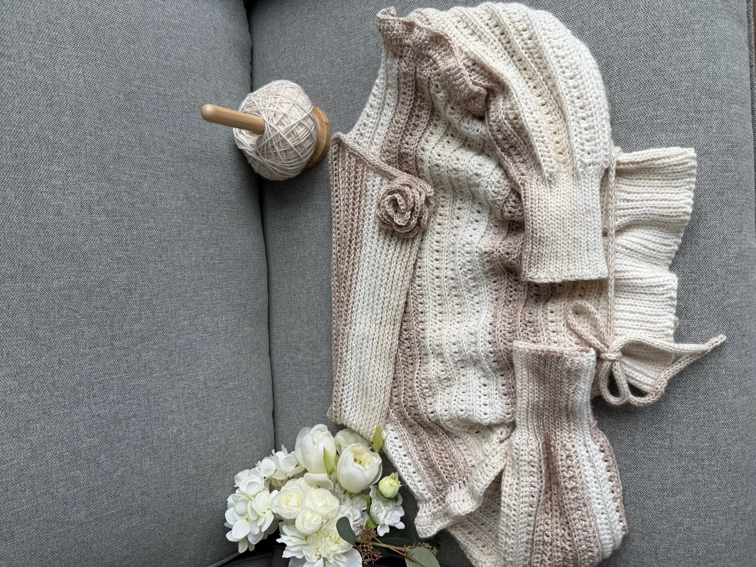 Knitted blanket, hat, and socks with a small wooden toy and white flowers on a gray surface.