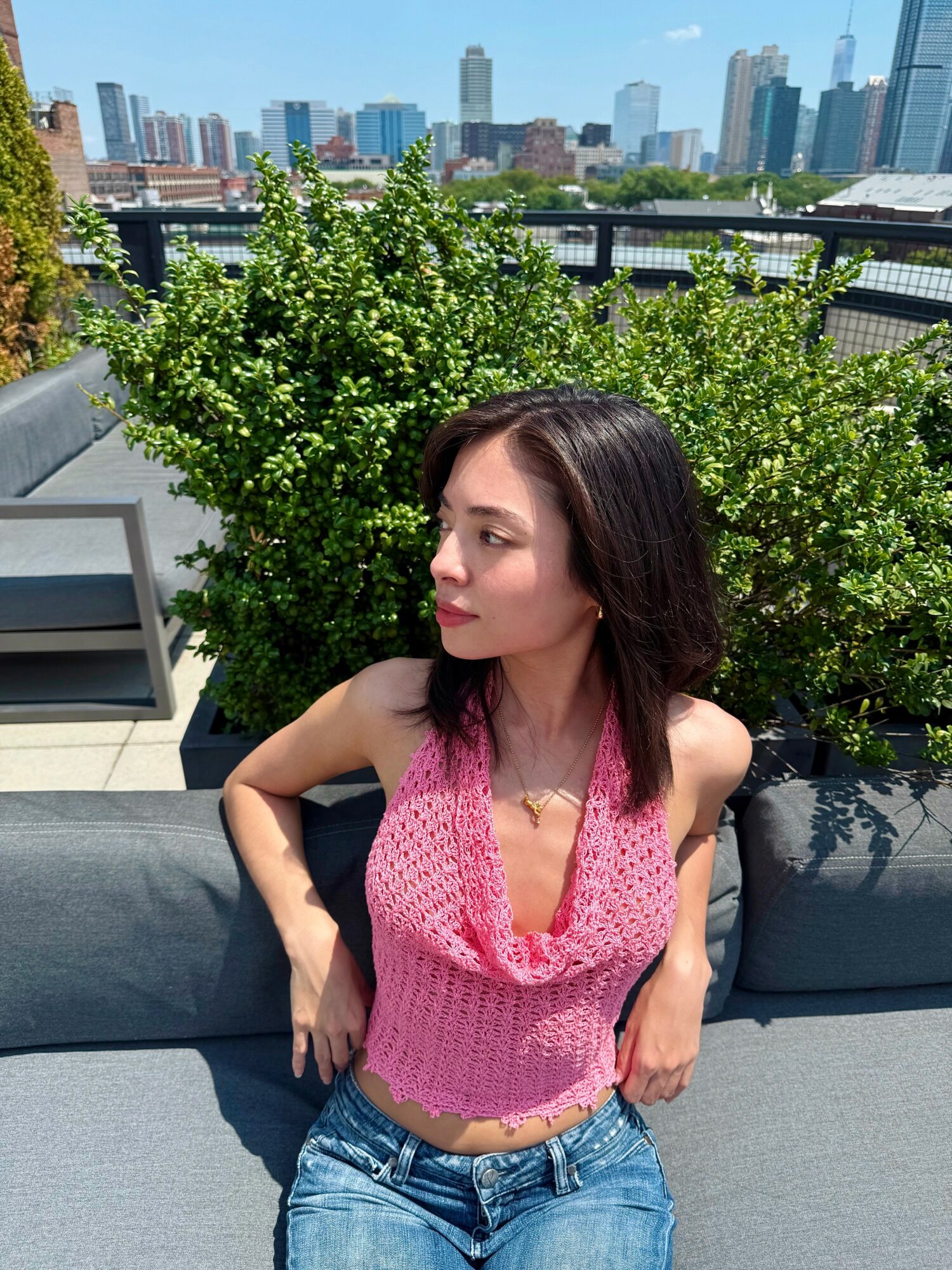 Woman with dark hair in pink sleeveless top and jeans sitting outdoors with city skyline in background.