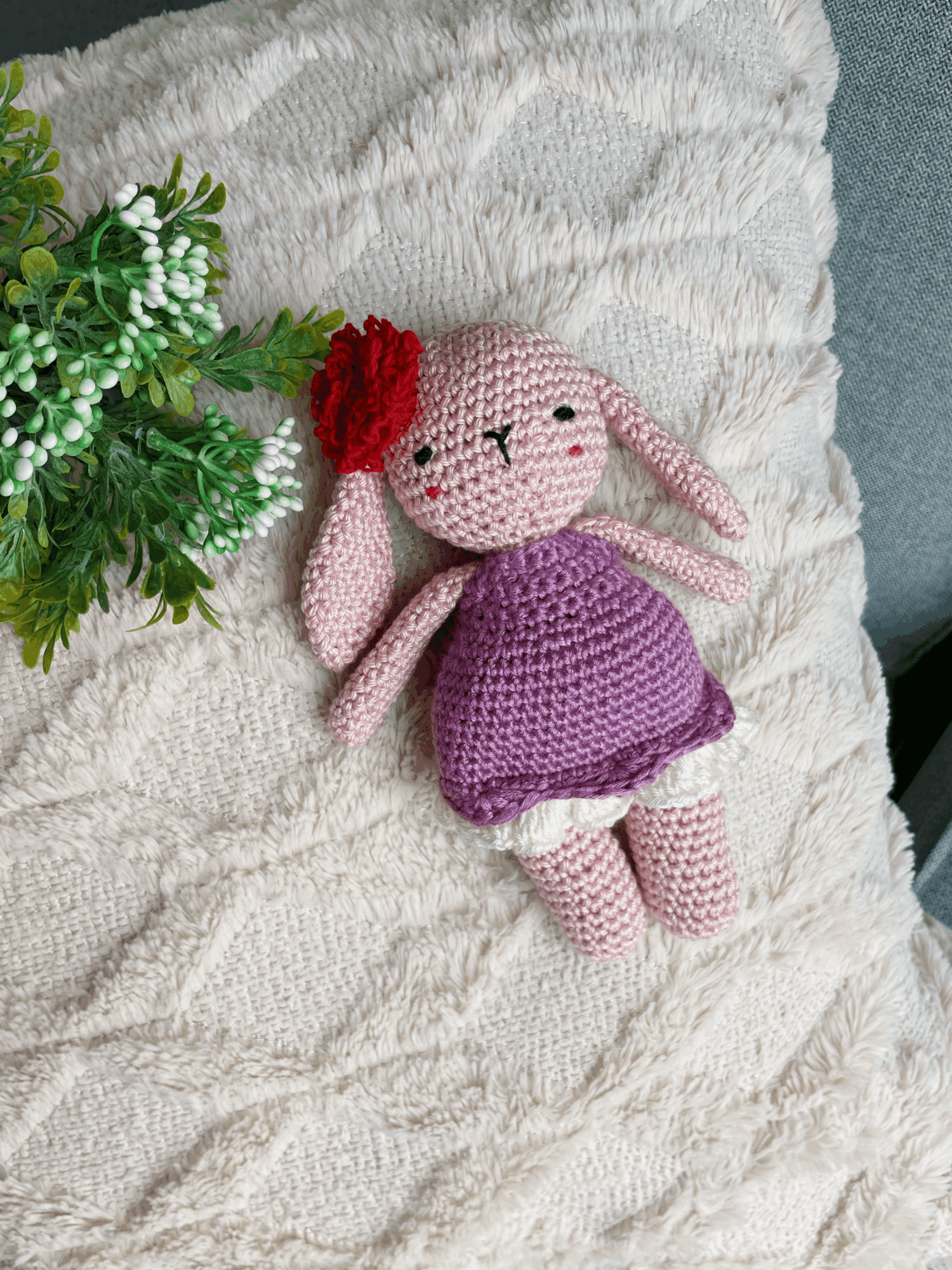 Pink knitted doll with red flower hair, purple dress, lying on a cream textured blanket, next to green plant leaves.