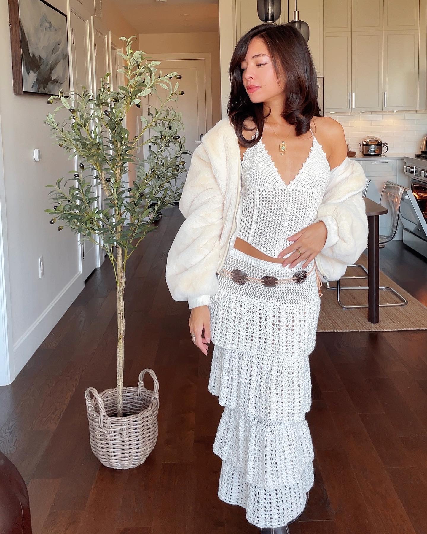 Woman in white dress and cardigan stands indoors near a potted plant, with a kitchen in the background.