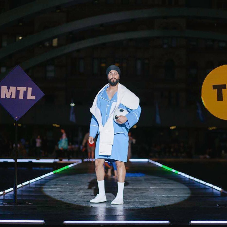 Man standing on a runway at night, wearing a blue and white jacket, shorts, and white shoes, with city buildings in background.