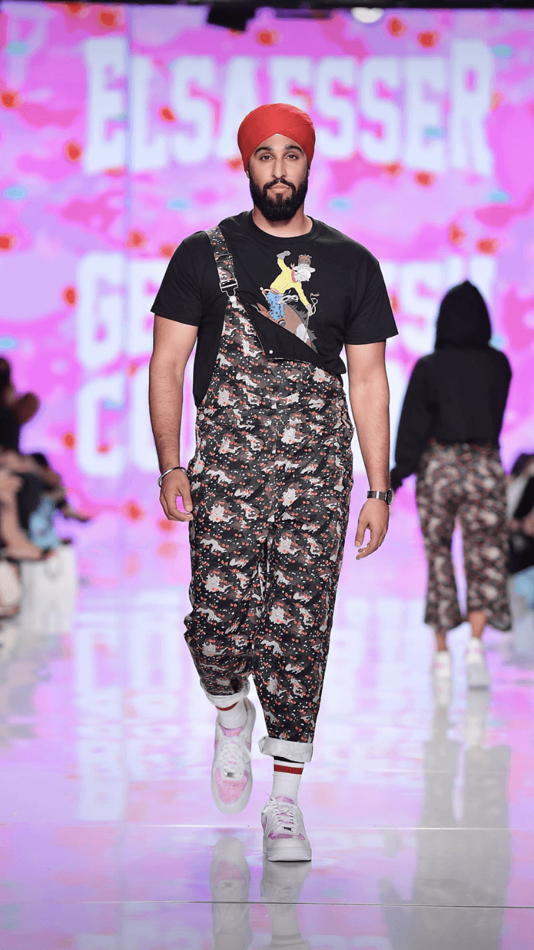 Male model walking on runway with pink background and text, wearing a red turban, black t-shirt, patterned pants, and white sneakers.