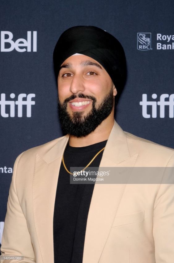 Man with a beard and black turban smiling, wearing a beige blazer and black shirt, standing in front of a dark background with logos.