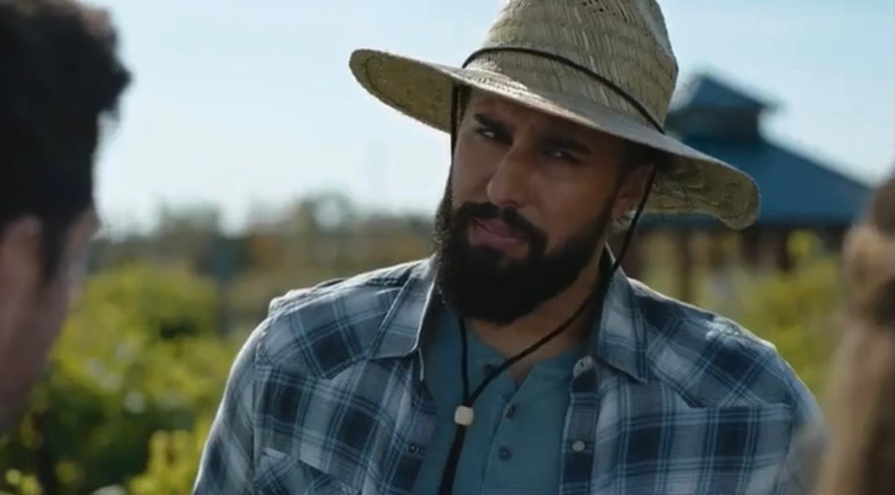 Man with a beard wearing a wide-brimmed hat and plaid shirt outdoors, talking to another person.