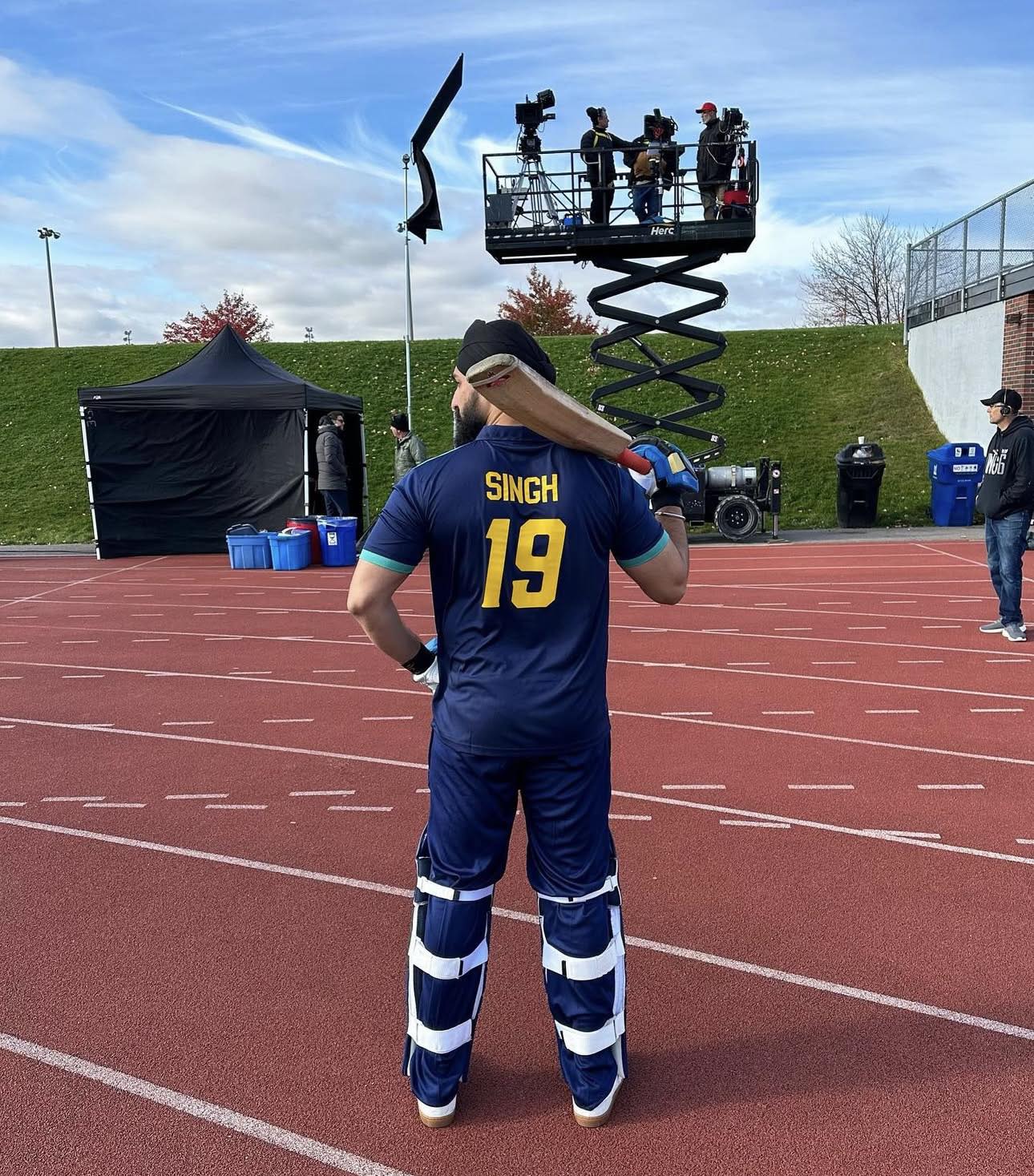 Person in a sports uniform with the name Singh and number 19, standing on a track, holding a bat, with camera crew above.