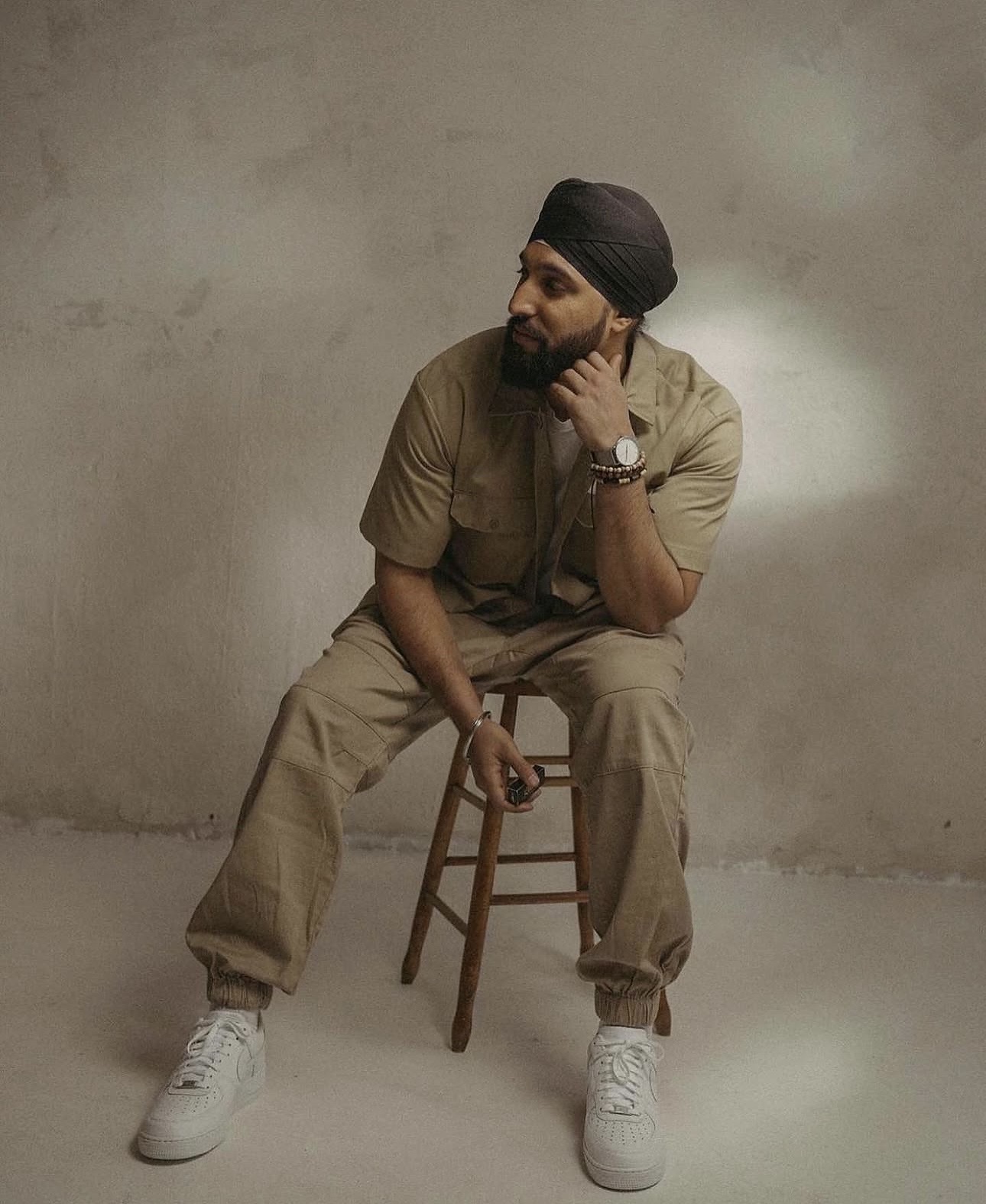 Man sitting on a wooden chair, wearing a turban, beige outfit, and white sneakers, against a plain wall.