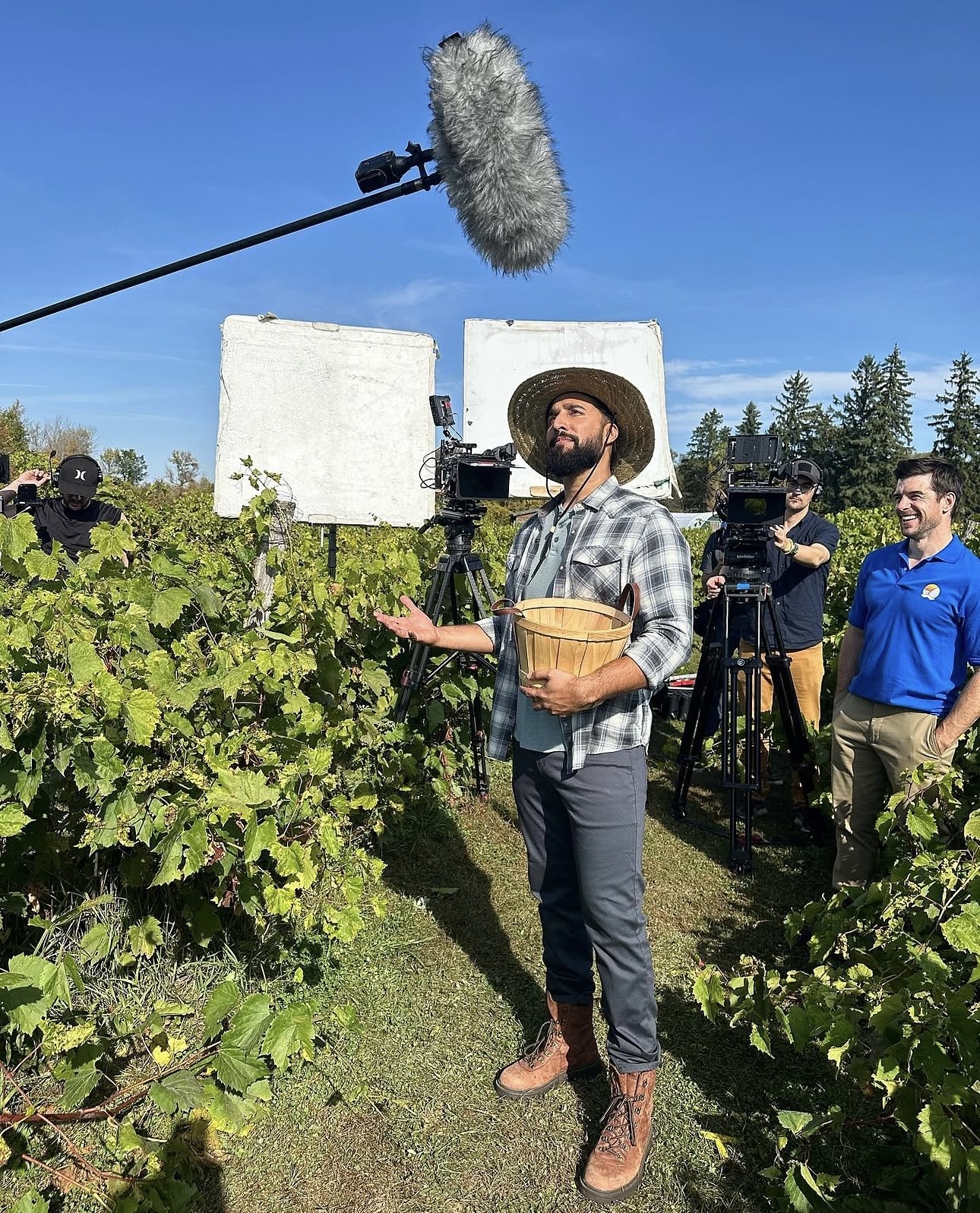 Filmmaker with camera and sound equipment in vineyard, crew members in background, clear blue sky, lush green vines.