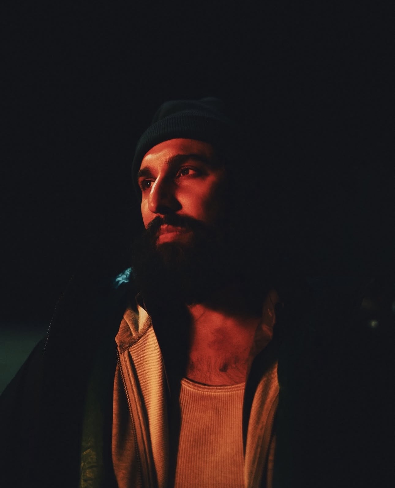 Portrait of a man with a beard, illuminated by warm light, wearing a beanie and jacket, against a dark background.