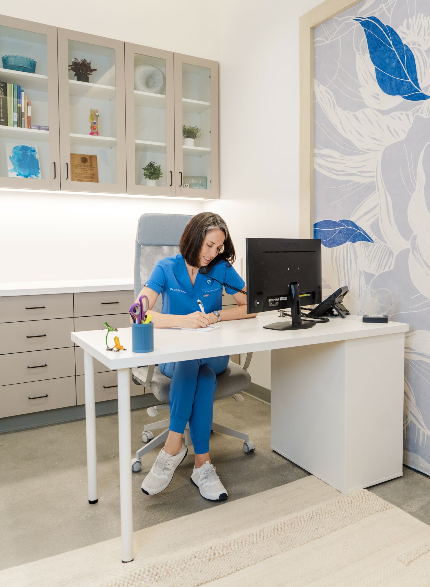 Woman in blue scrubs sitting at a white desk with a computer, in a modern office with cabinets and a large wall art.