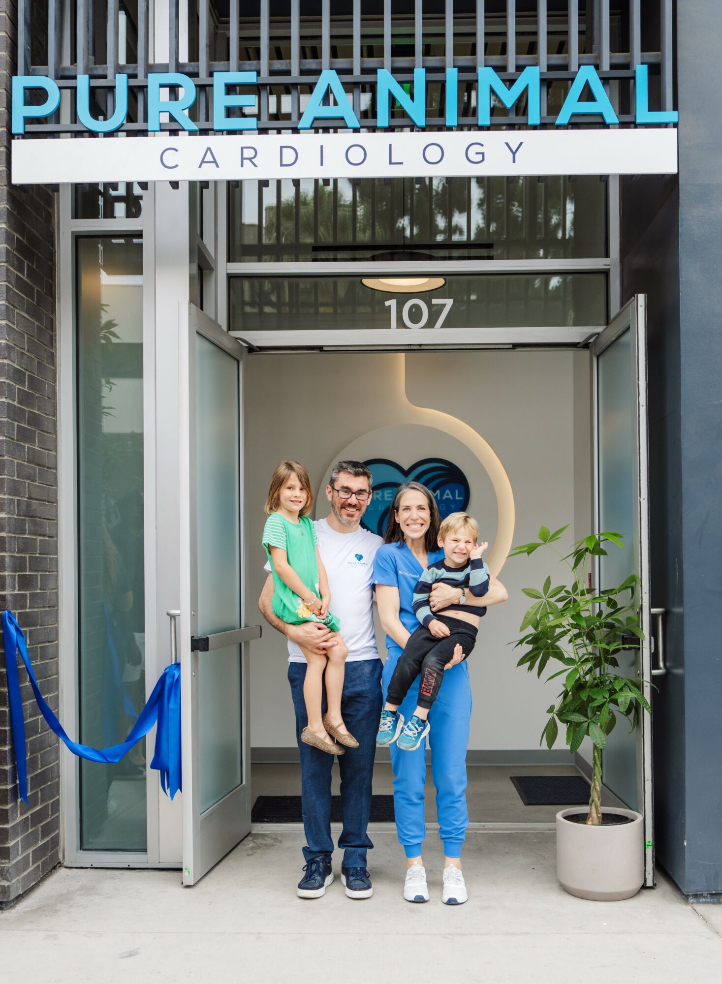 Group of four people standing outside a clinic entrance with a sign reading 'PURE ANIMAL CARDIOLOGY', smiling, with a potted plant nearby.