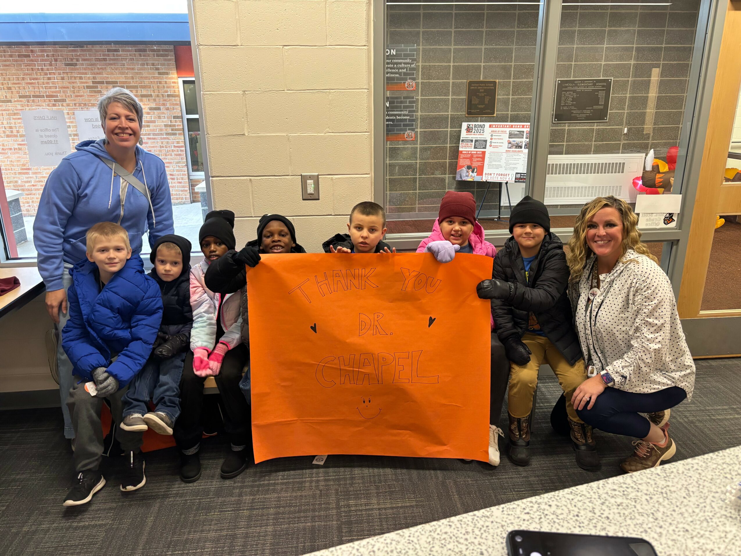 Group of children and two adults holding a large orange poster board inside a room with windows and a counter.