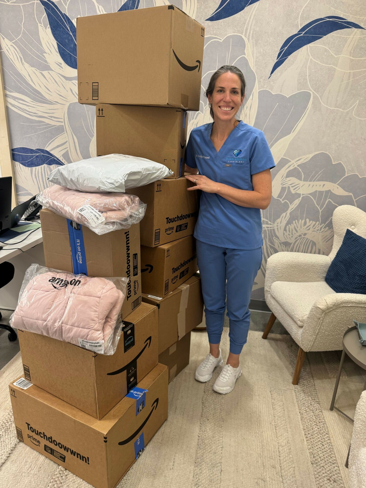 Person in blue scrubs standing next to stacked boxes and packages in a room with floral wallpaper.
