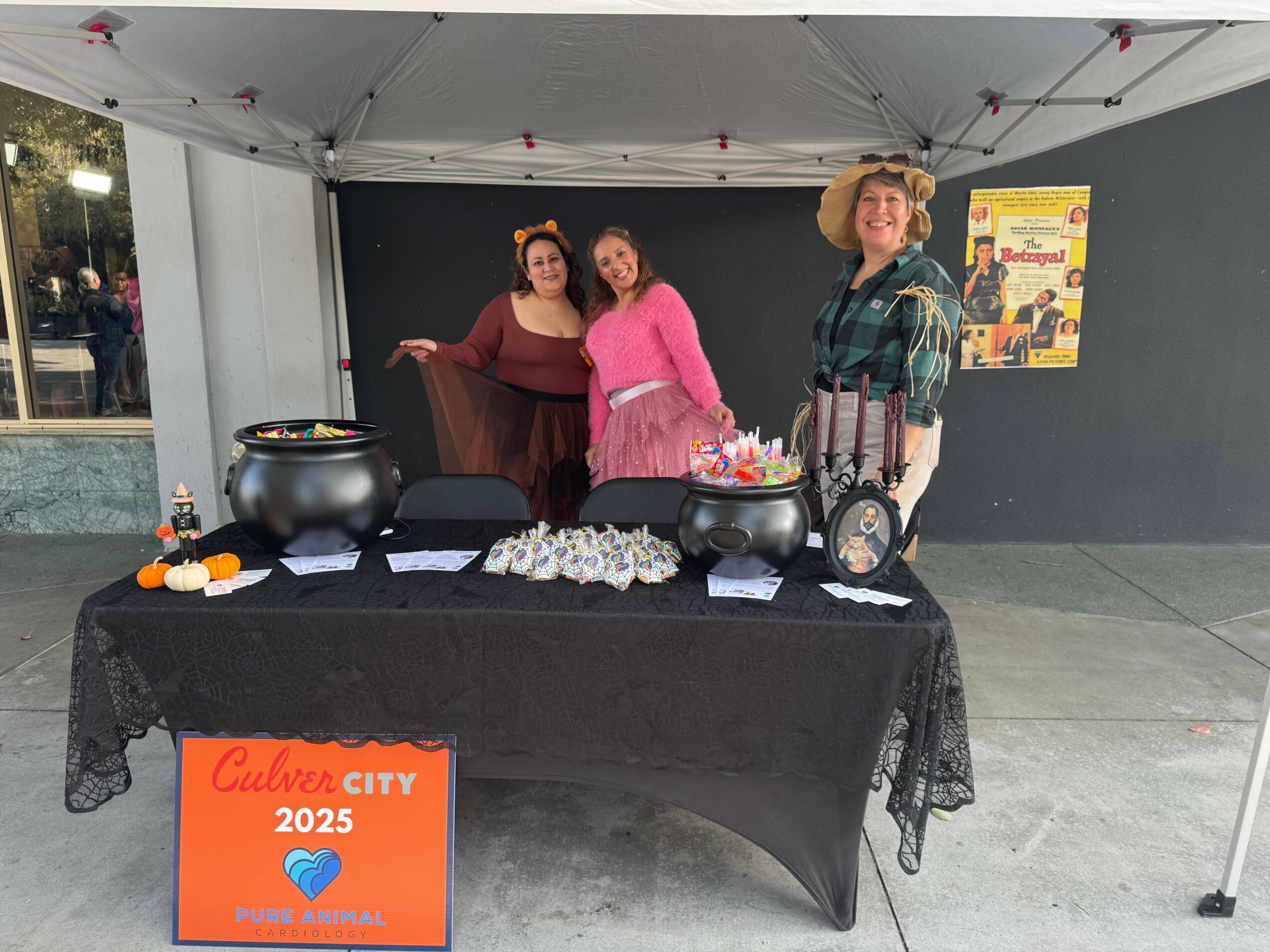 Three women stand behind a table with food and decorations under a canopy, with a poster on the wall behind them.