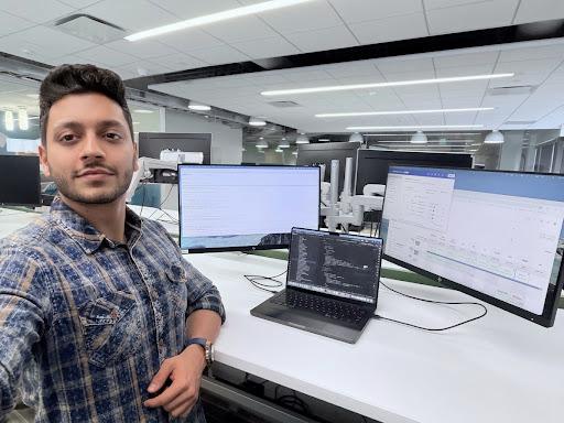 Man in a plaid shirt in an office with multiple computer monitors and a laptop, looking at the camera.