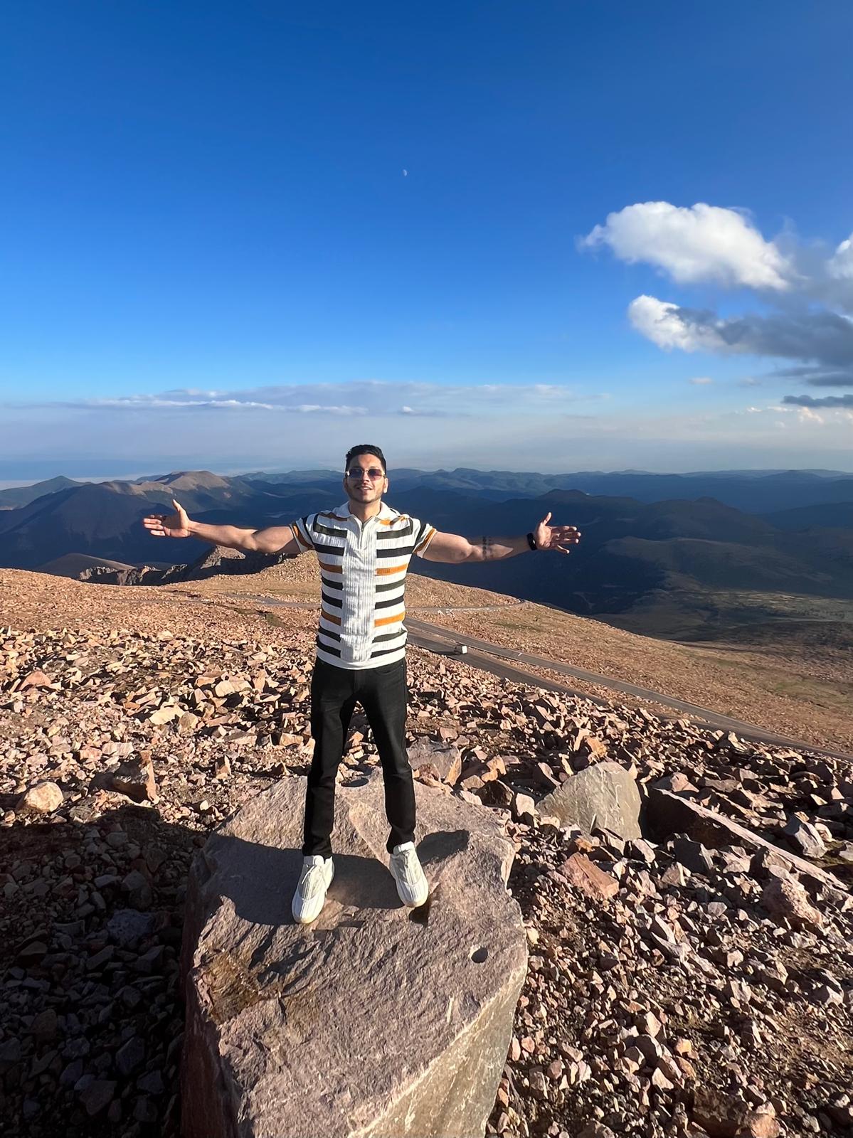 Person standing on rocks with arms outstretched, mountain landscape and blue sky in background.