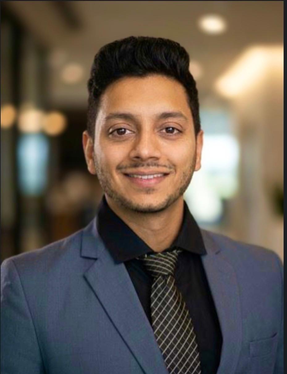 Man in a suit smiling, indoor background with blurred lights and windows.