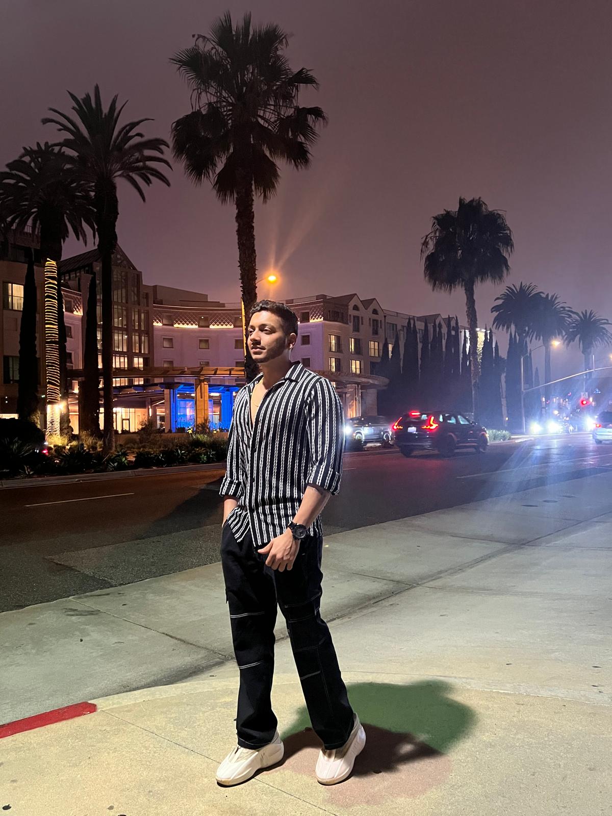 Person standing on sidewalk at night with palm trees and city buildings in background.