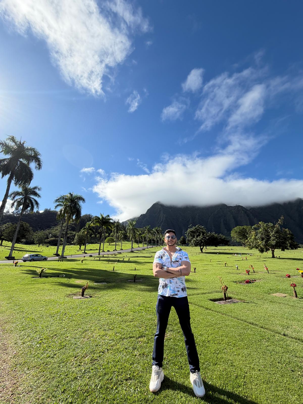 Person standing on a grassy field with mountains and palm trees in the background under a blue sky with clouds.