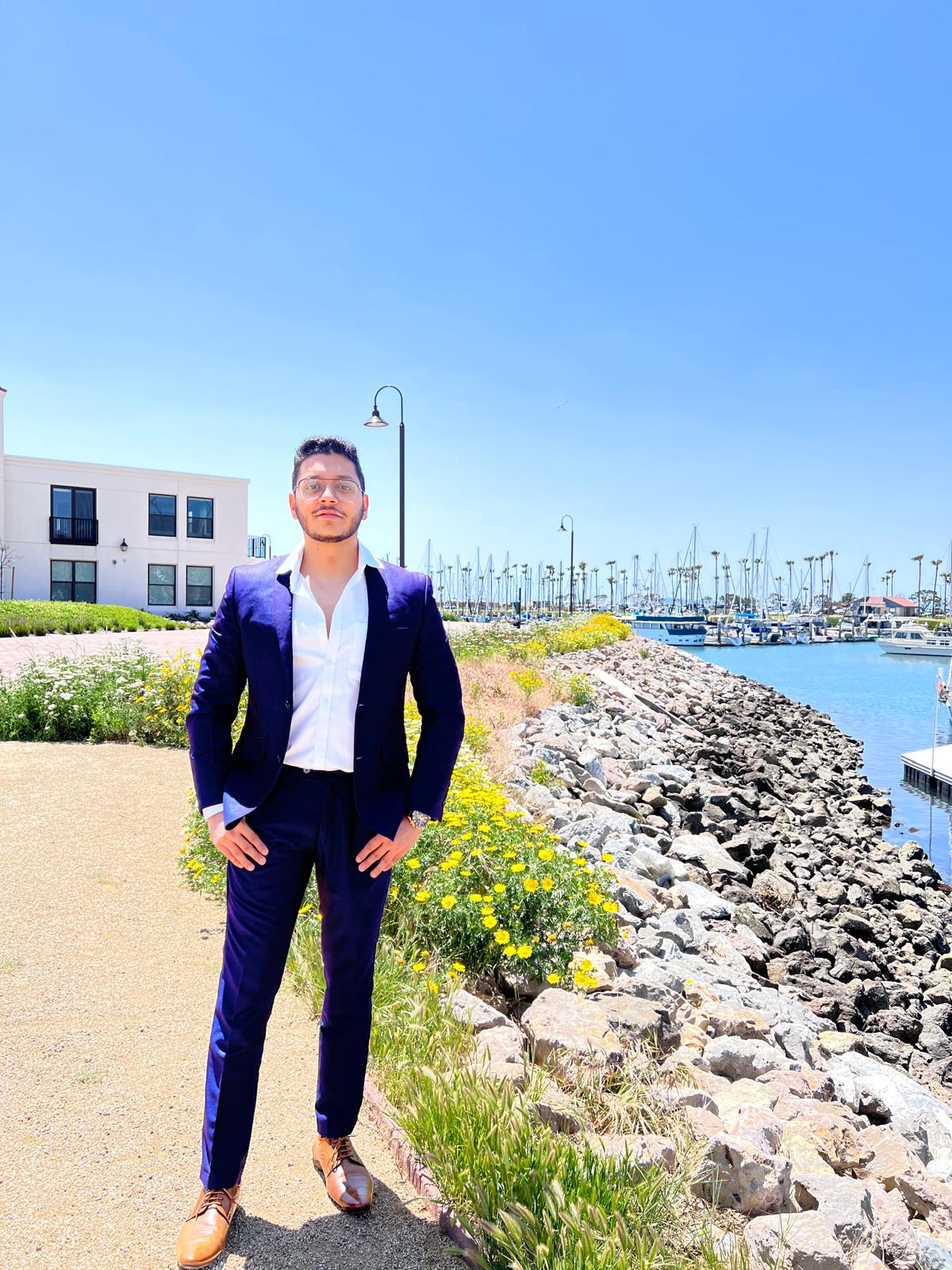 Man in a navy suit standing on a path near a marina with boats, buildings, and a clear blue sky in the background.