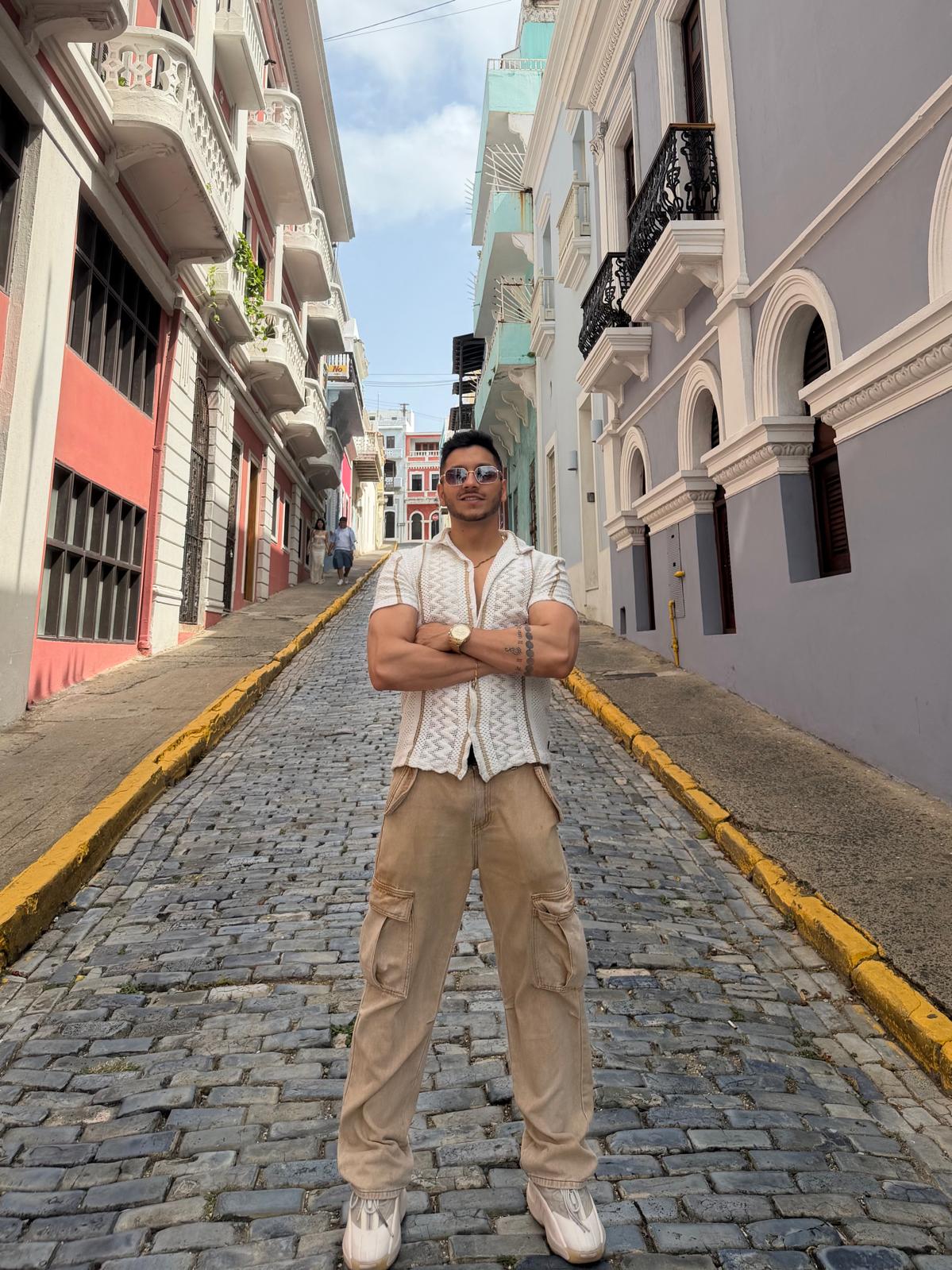 Young man standing with arms crossed on cobblestone street, surrounded by colorful buildings with balconies and decorative architecture.