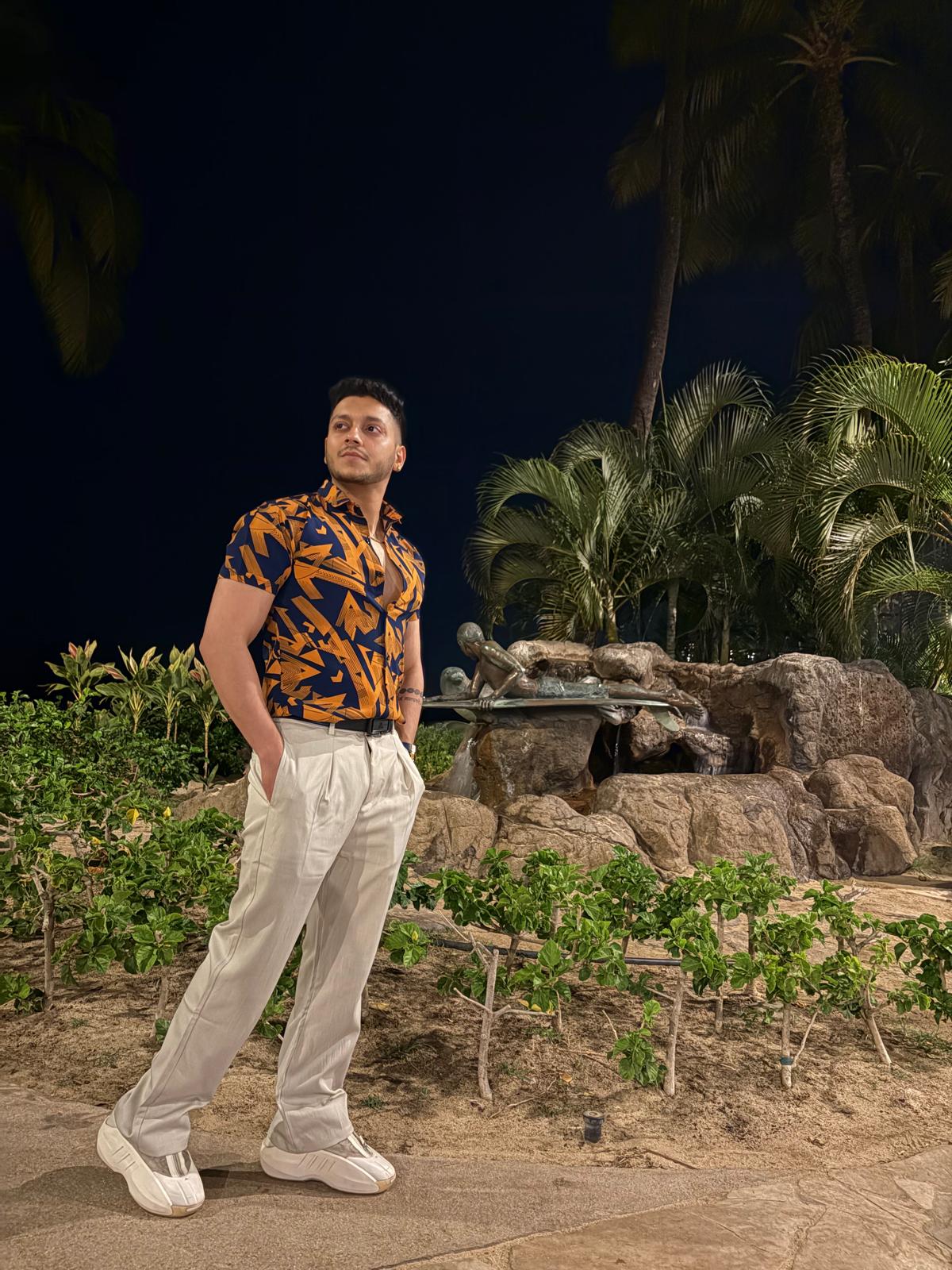 Young man standing outdoors at night, surrounded by plants and rocks, wearing a patterned shirt and light-colored pants.