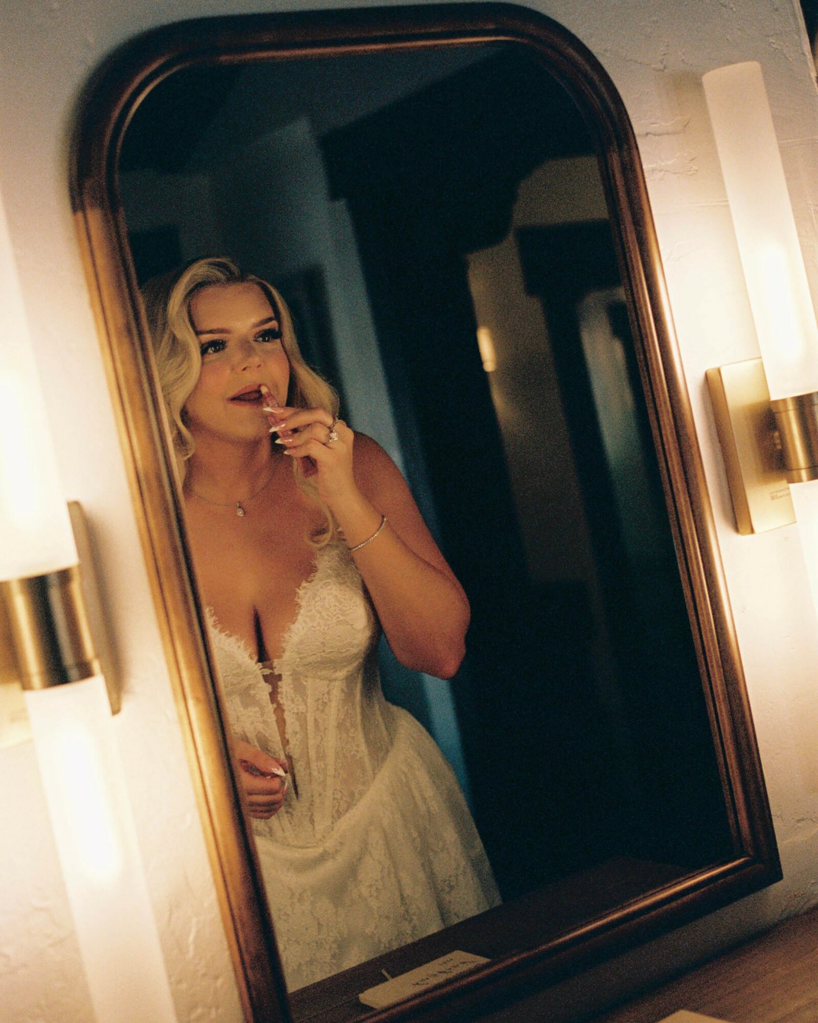 Woman in a white dress looking in a mirror, holding a cigarette, illuminated by wall lights, with a dark background.