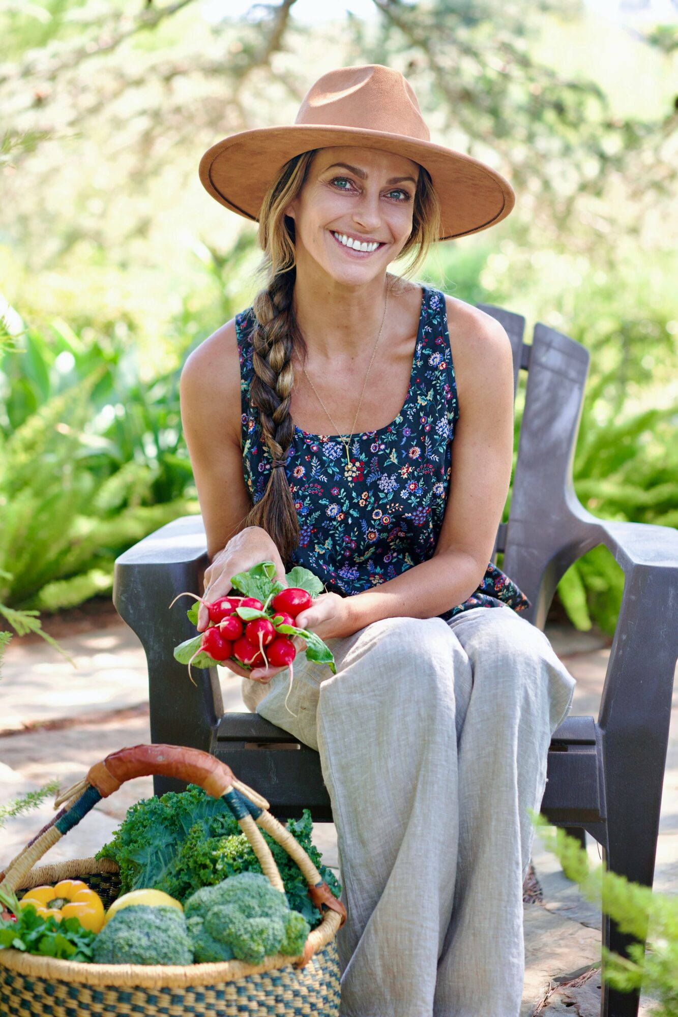 Woman sitting outdoors on a bench, holding a bouquet of flowers, wearing a hat and floral top, smiling.