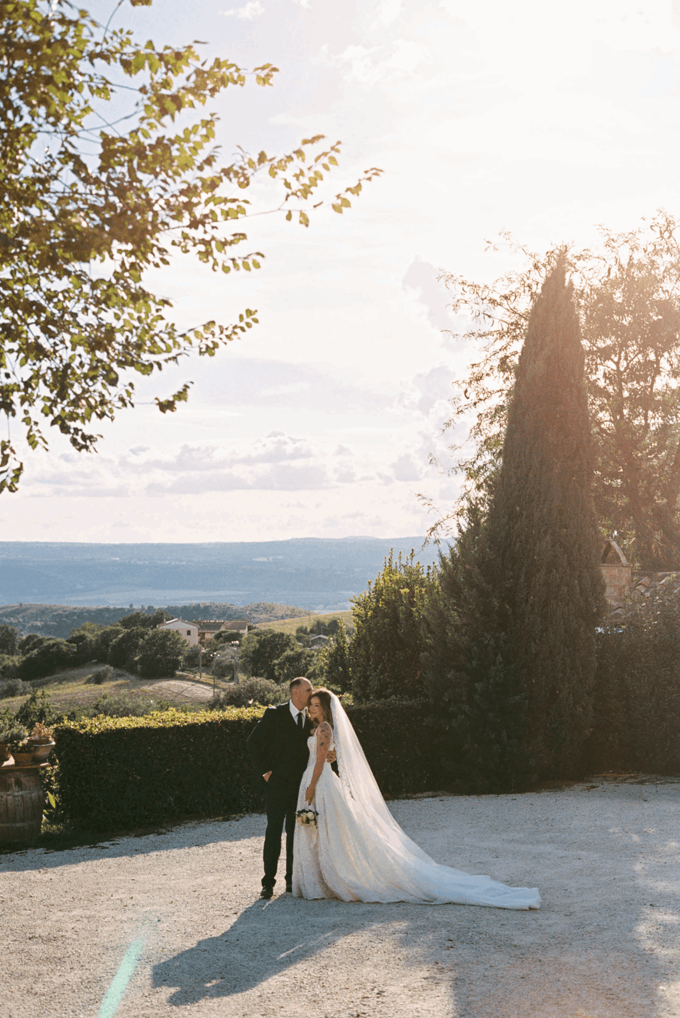 A bride and groom stand together outdoors with a scenic landscape in the background, trees, and a cloudy sky.