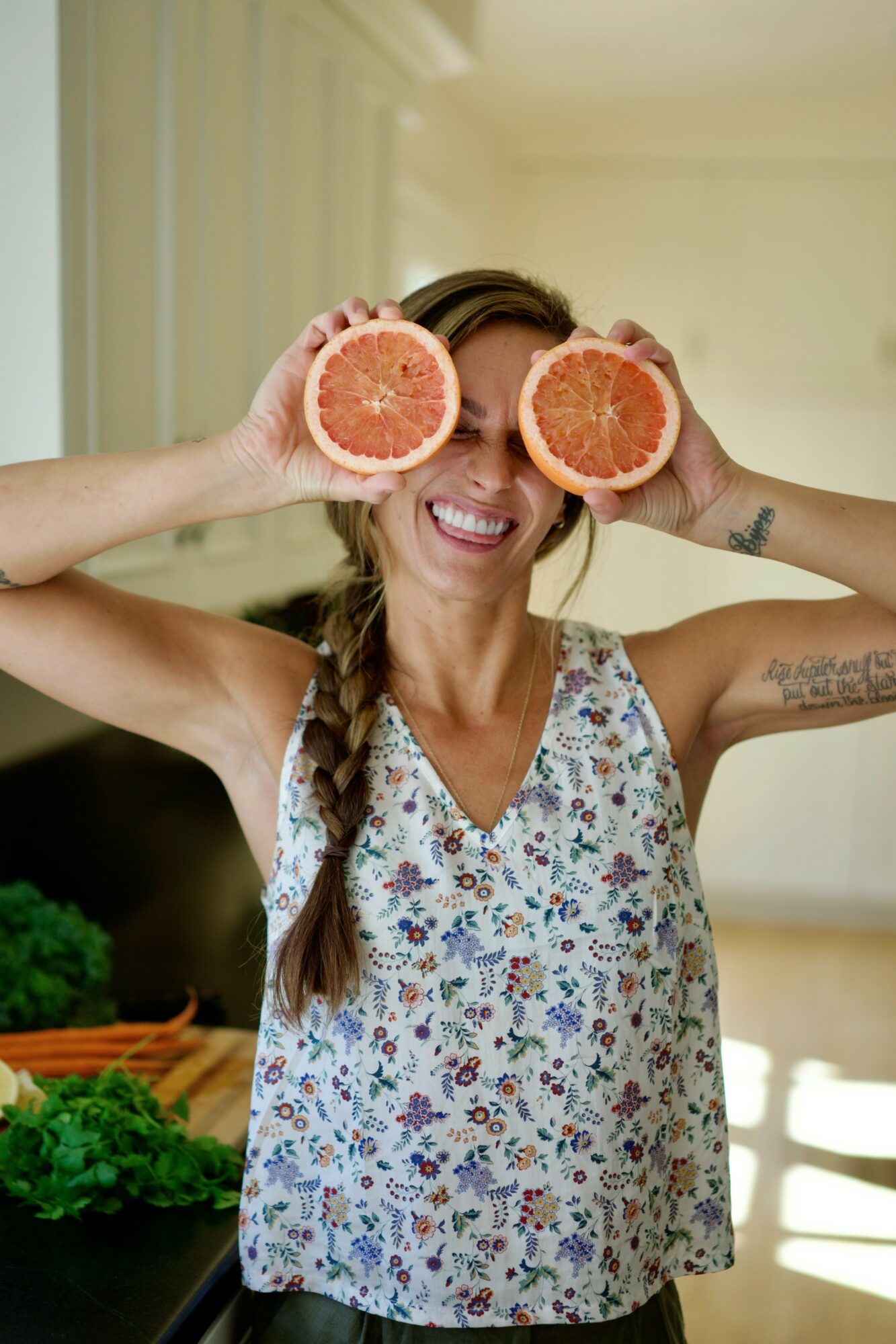 Woman holding grapefruit halves in front of her eyes, smiling, in a bright room with plants and sunlight.