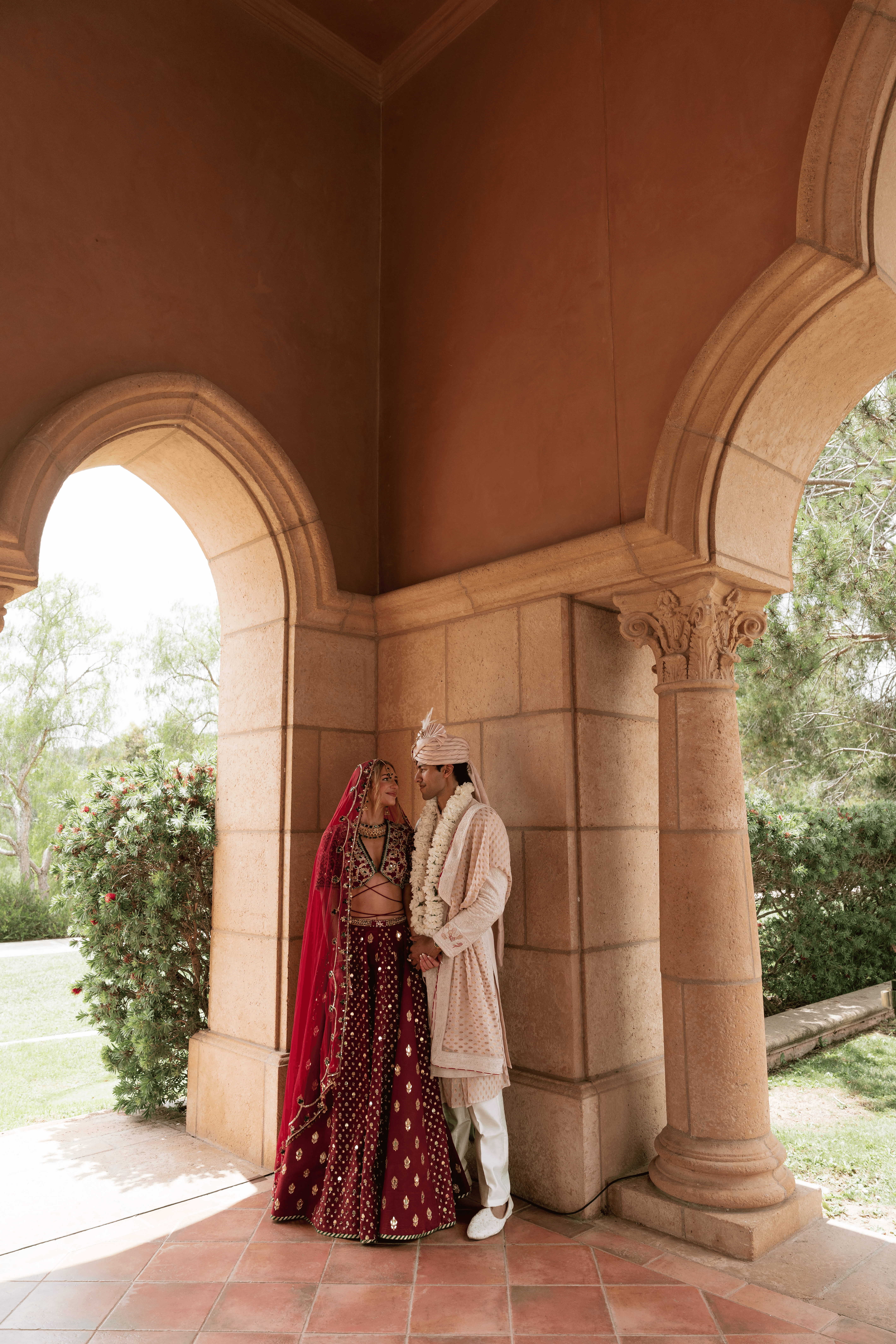 Two people in traditional attire stand under an arched stone structure with columns, greenery visible outside.