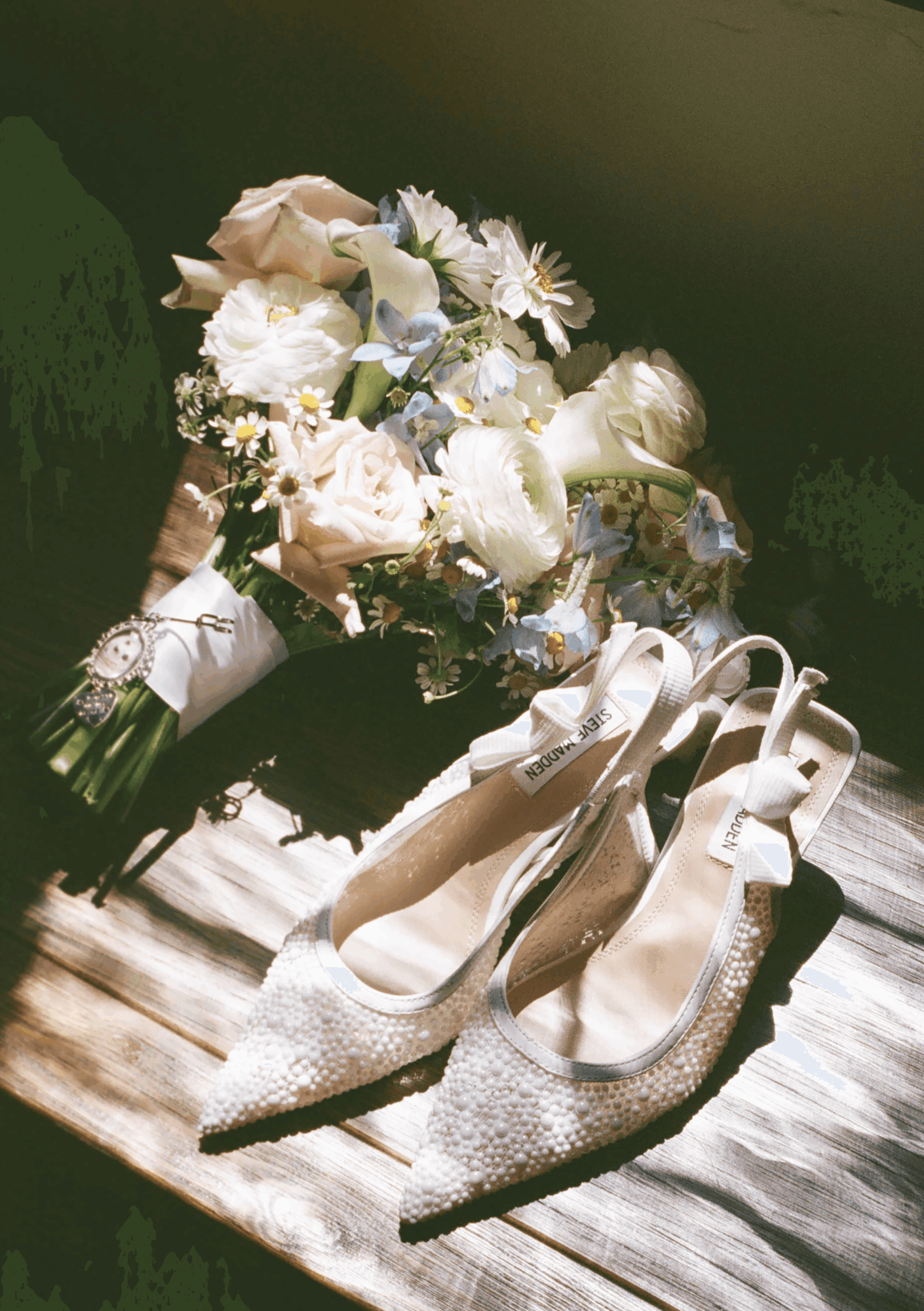 A bouquet of white and pale pink flowers with ribbons, placed next to a pair of glittery high-heeled shoes on a wooden surface.