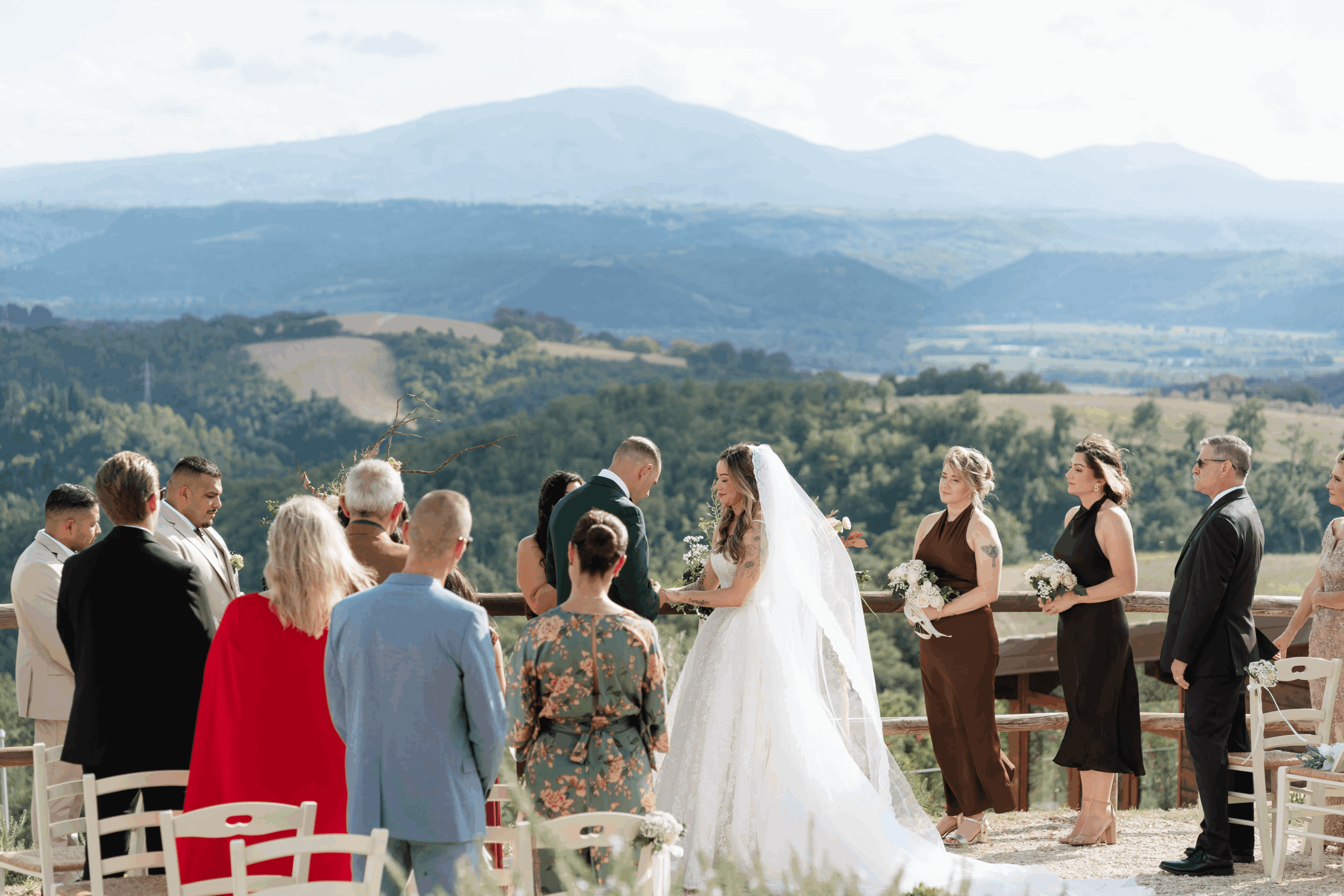 A wedding ceremony outdoors with a bride in a white dress and veil, surrounded by guests, against a scenic landscape background.