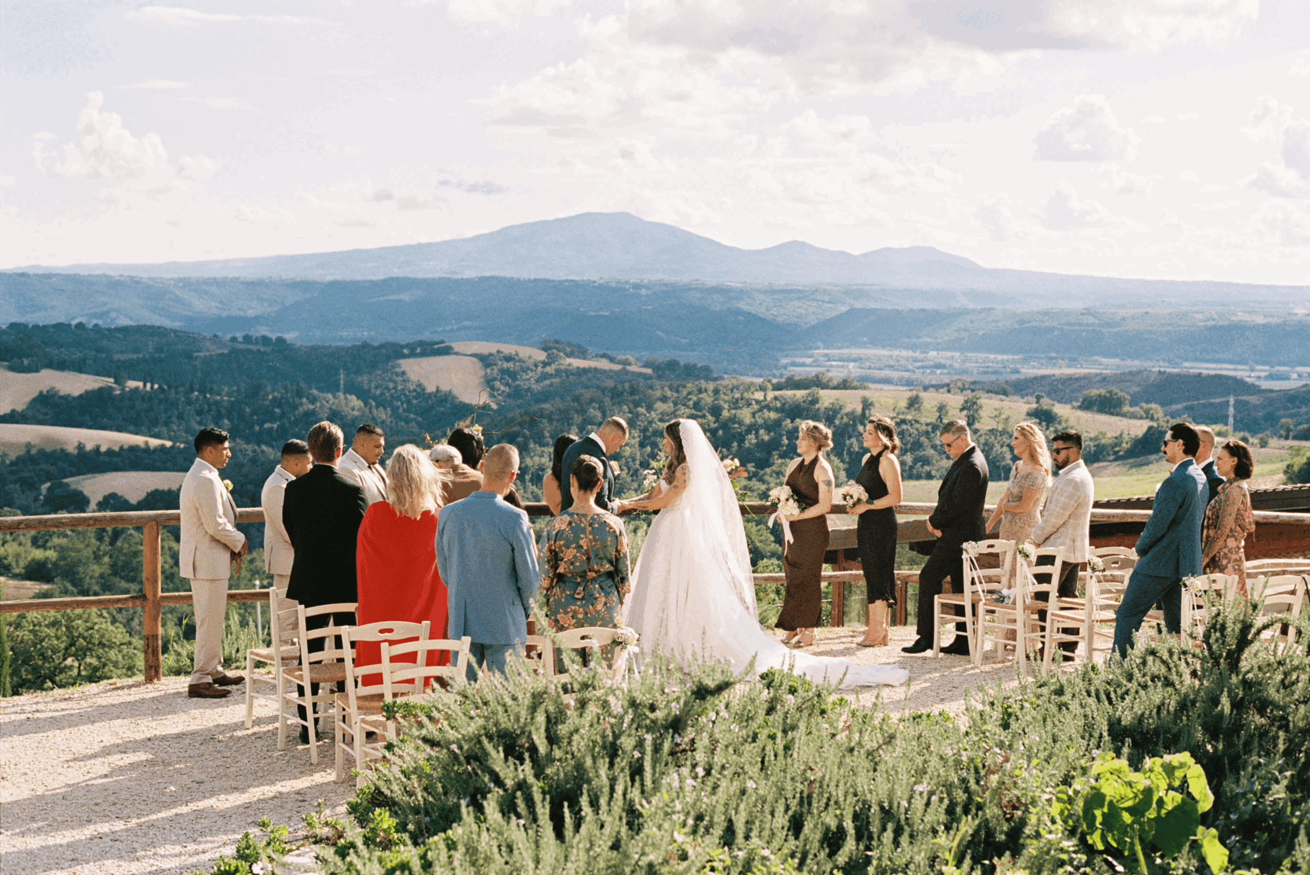 Wedding ceremony outdoors with people, bride in white dress, scenic landscape background, chairs arranged for the event.