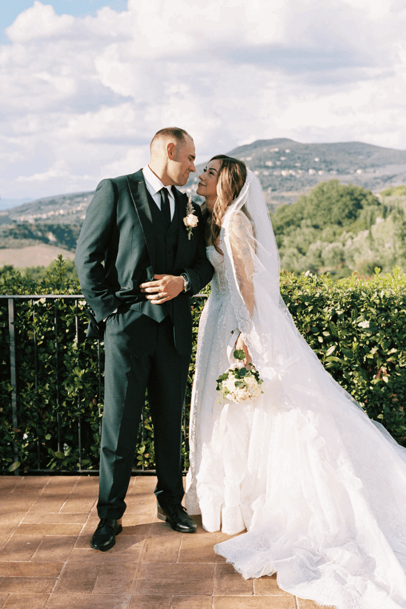 Bride and groom standing outdoors, facing each other, with scenic hills and sky in background.