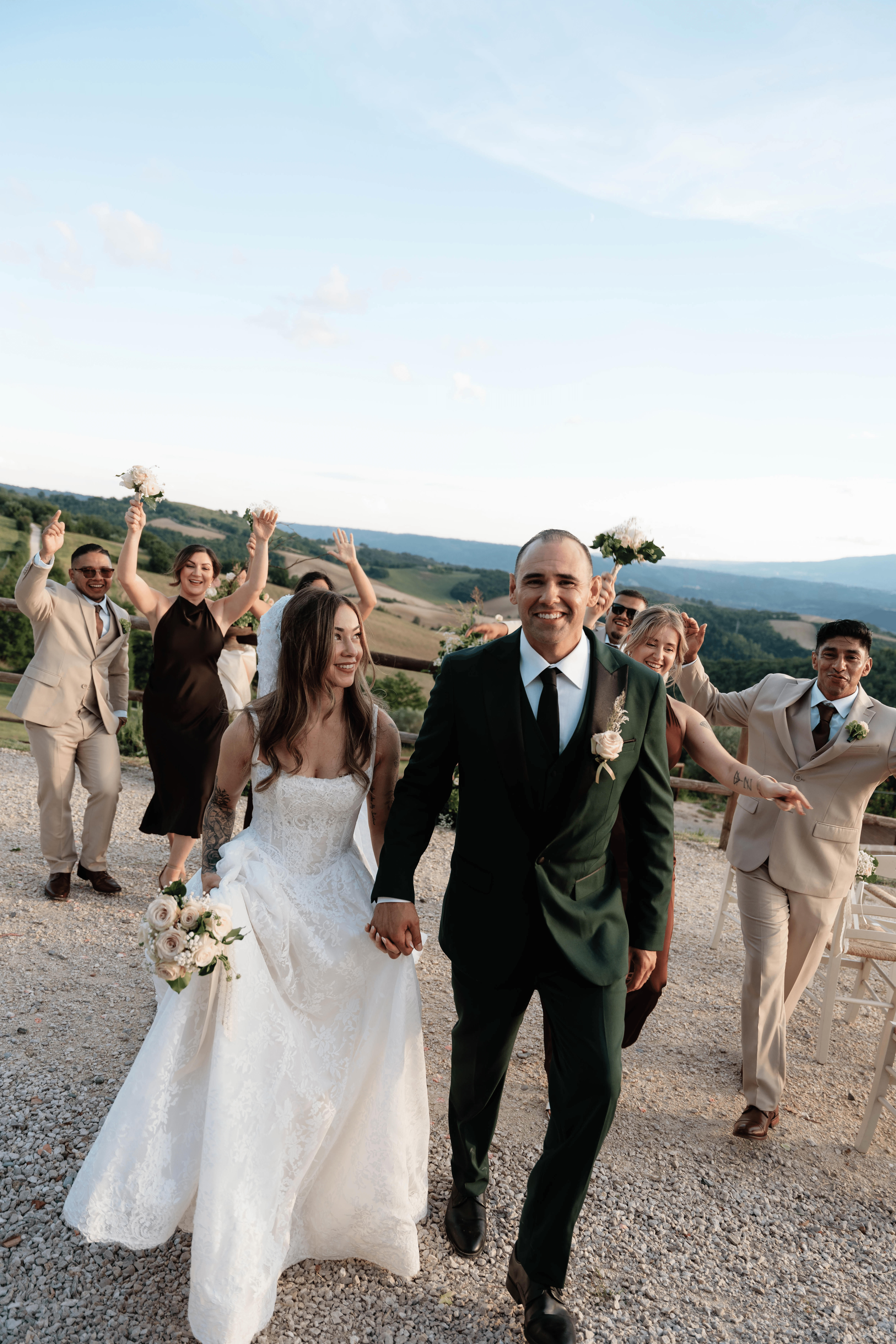 A wedding couple walking outdoors with friends celebrating behind them, scenic landscape in the background, sunny day.