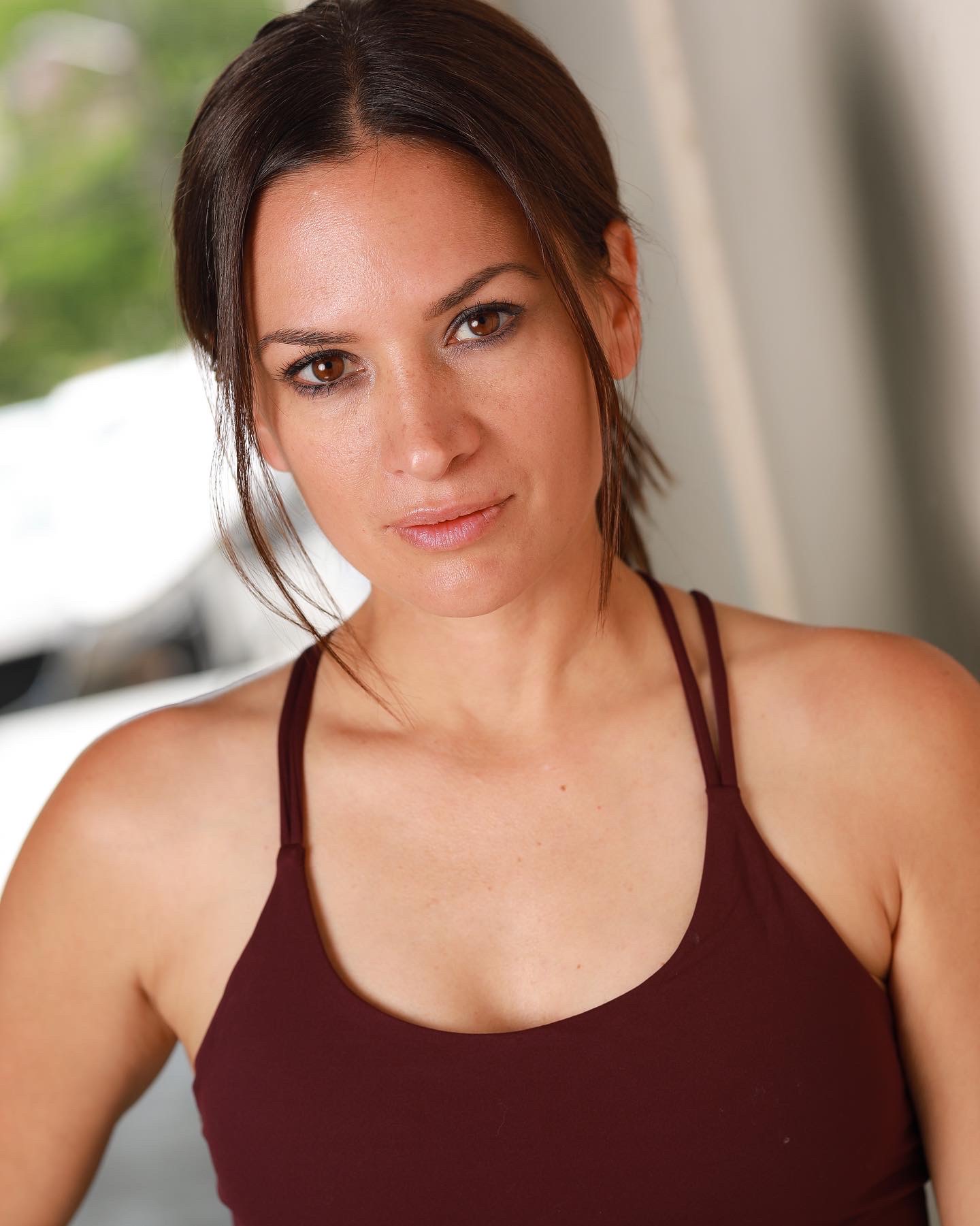 Woman with dark hair in a ponytail wearing a maroon tank top, looking at the camera indoors.