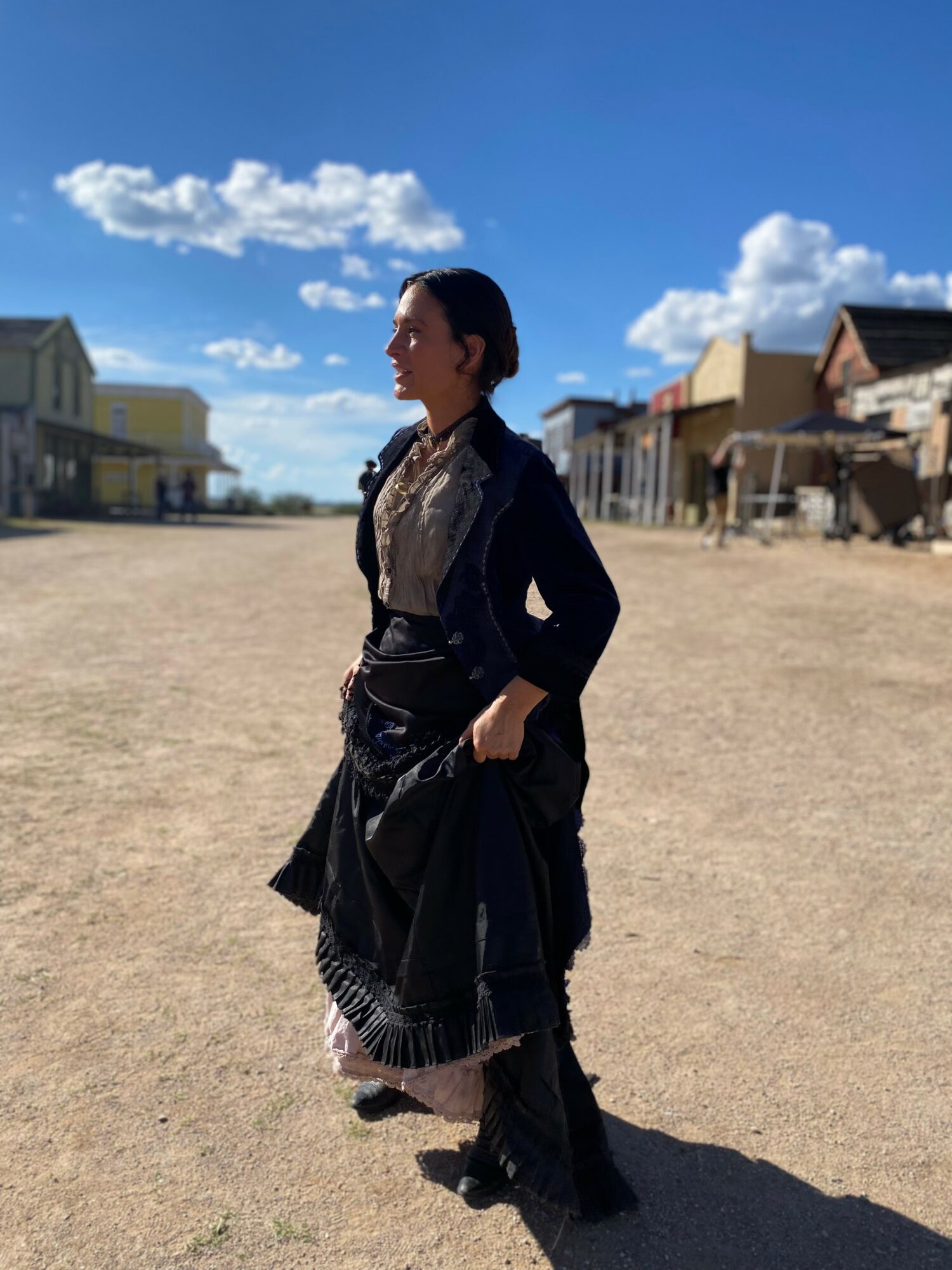 Woman standing outdoors in a western-style town, wearing a dark jacket and skirt, with buildings and a blue sky in background.
