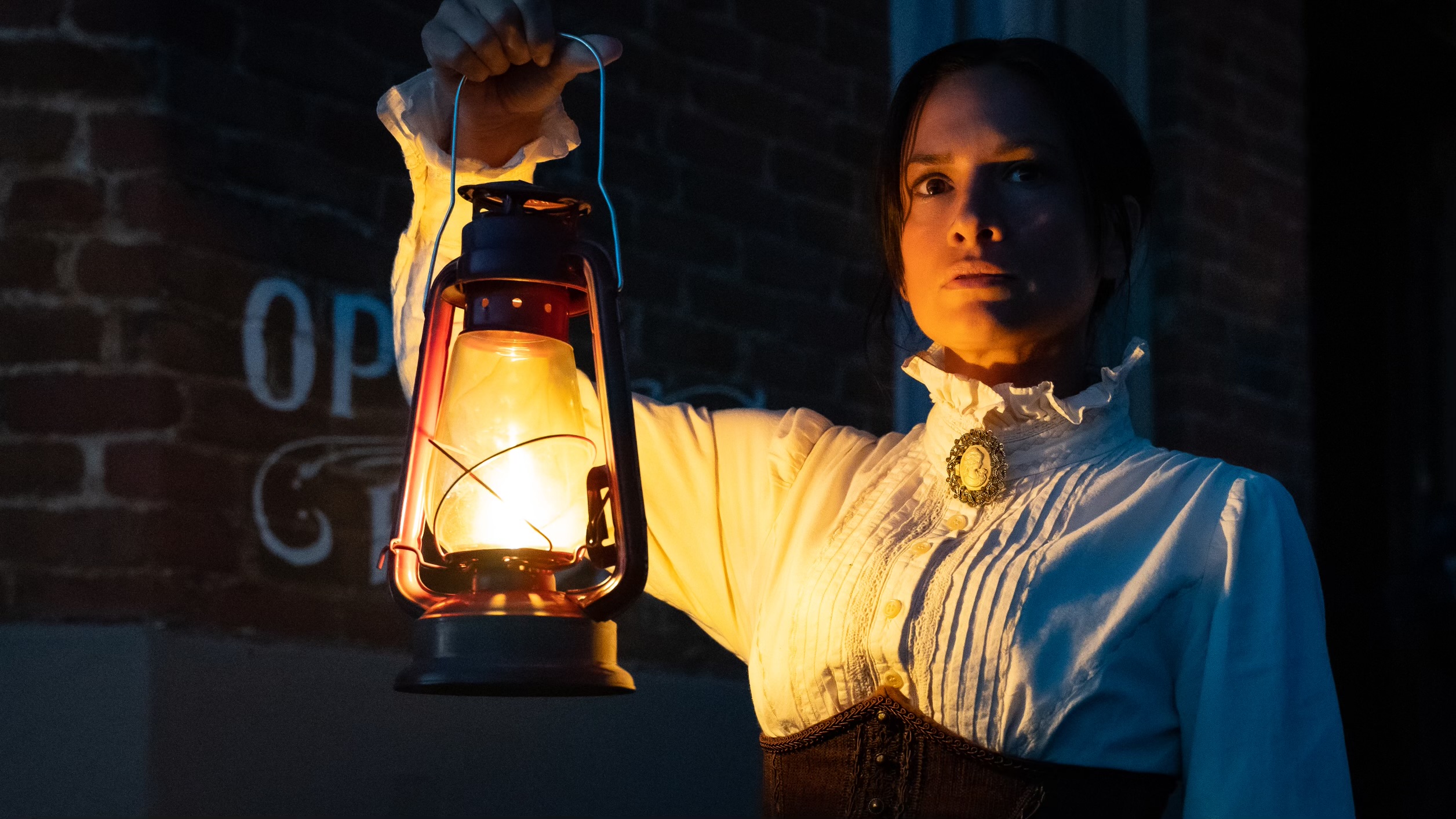 Woman holding a lit lantern in front of a brick wall at night, with a serious expression.