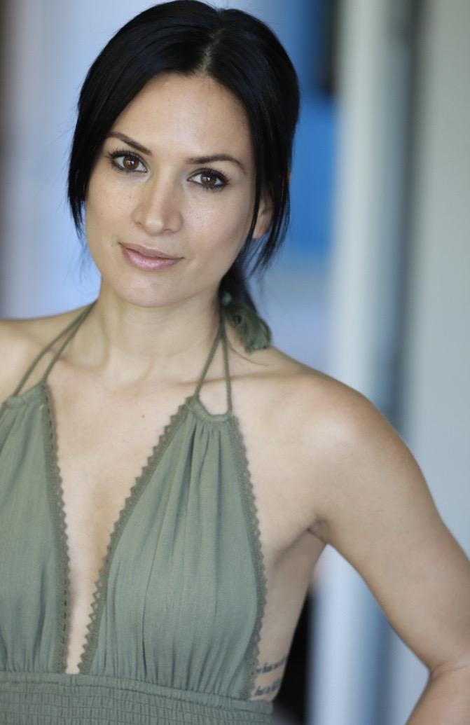 Woman with dark hair in a green halter top, standing indoors with a blurred background.